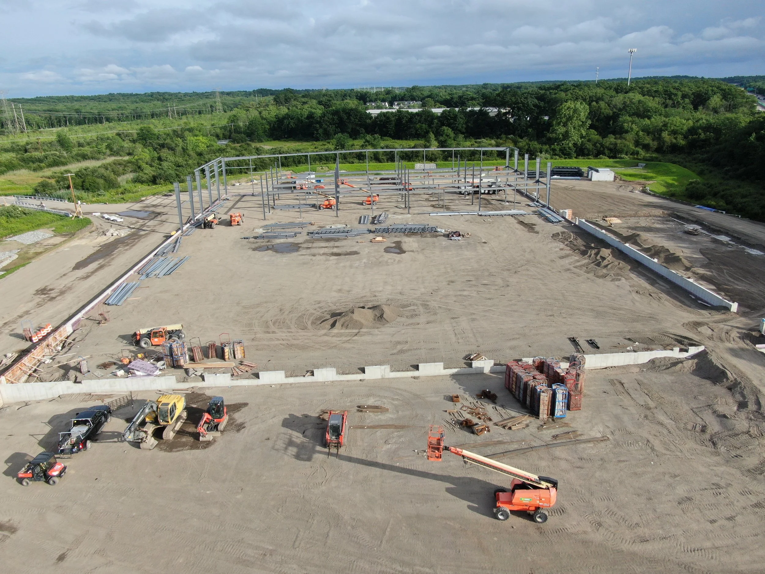 A construction site with a steel framework being assembled, surrounded by construction vehicles and equipment, with a green, hilly landscape and cloudy sky in the background.
