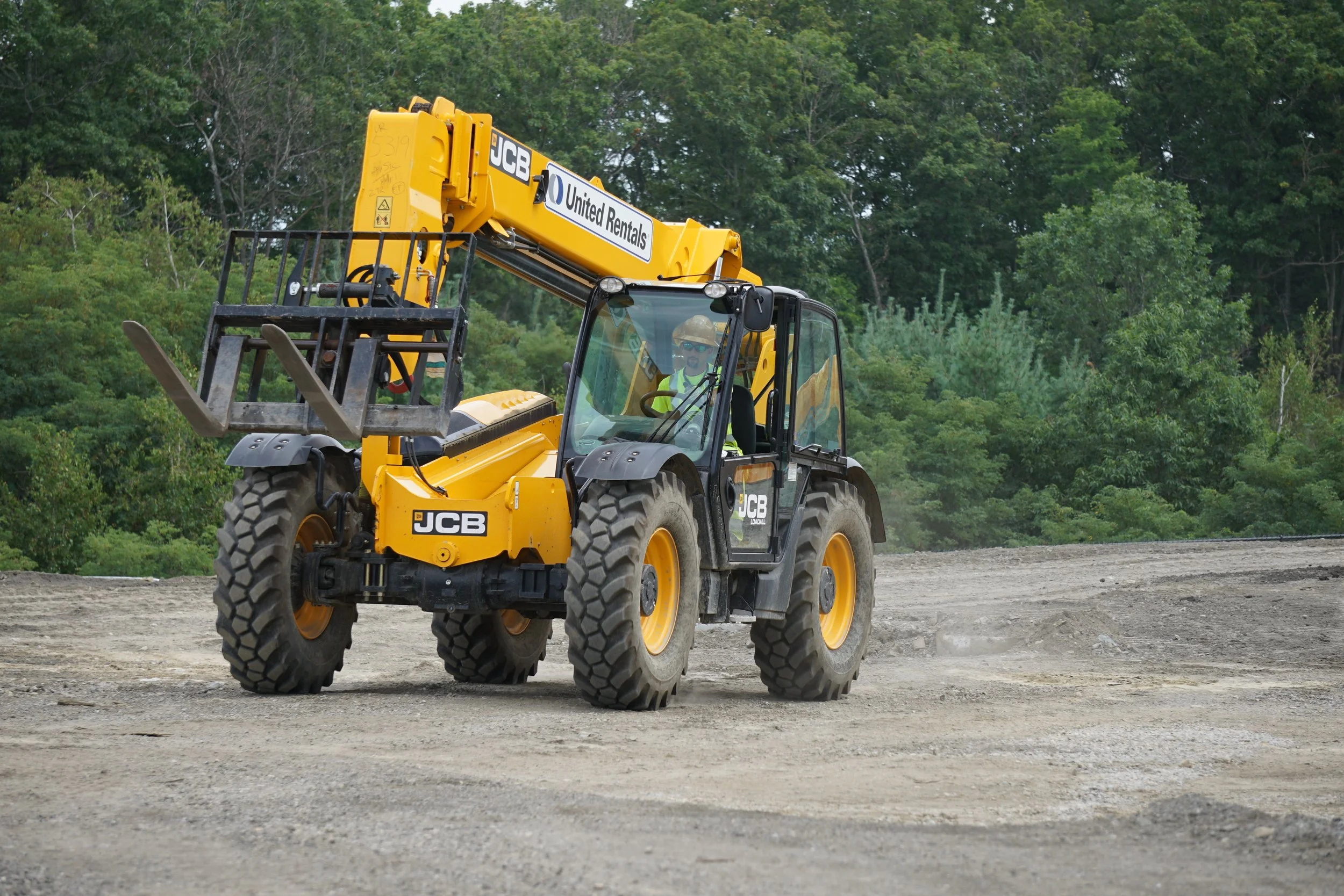 A yellow JCB telehandler at a construction site with a worker inside the cabin, surrounded by trees.