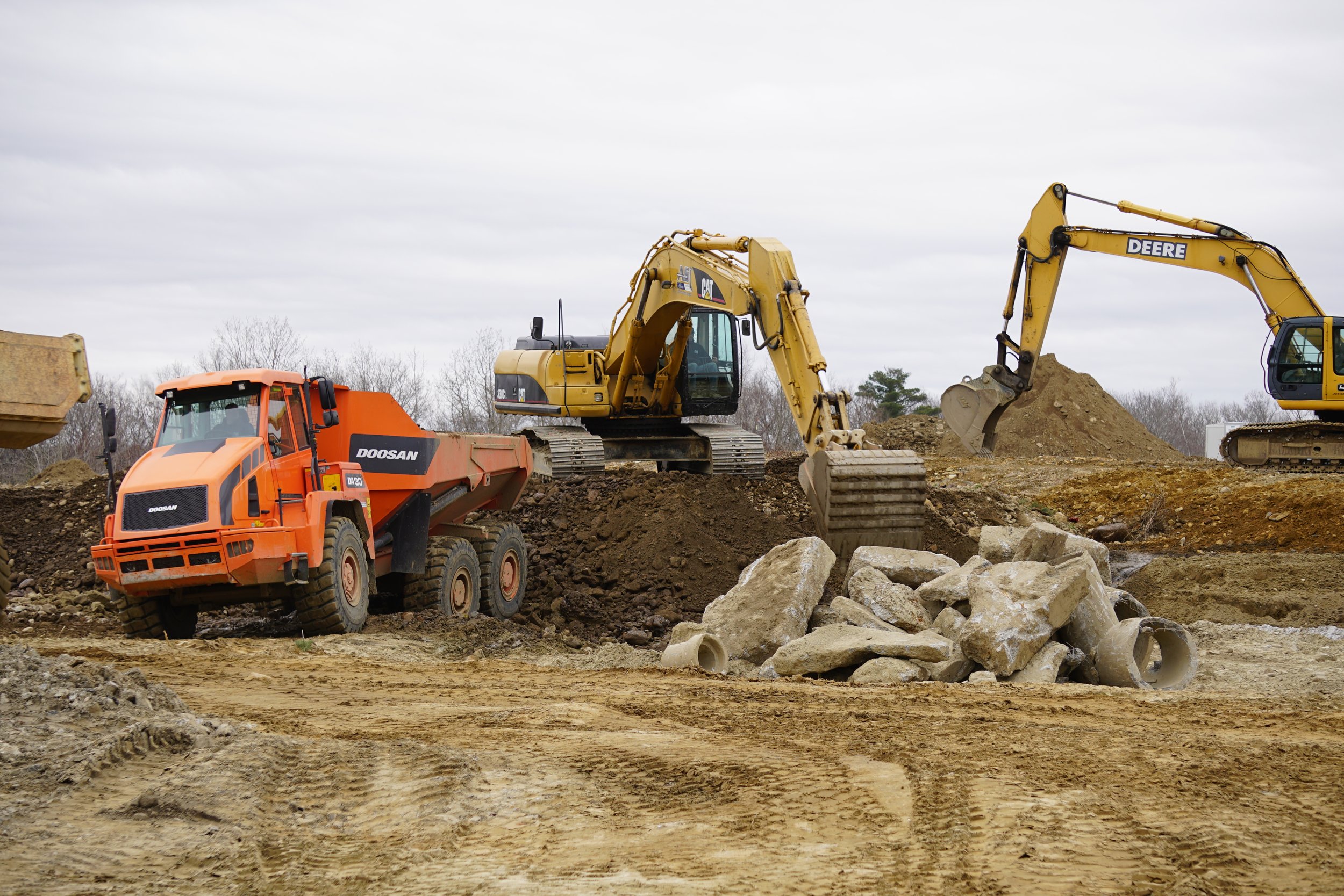 Construction site with heavy machinery including an orange road roller, a yellow excavator, and a yellow backhoe loader working on dirt and rocks under an overcast sky.