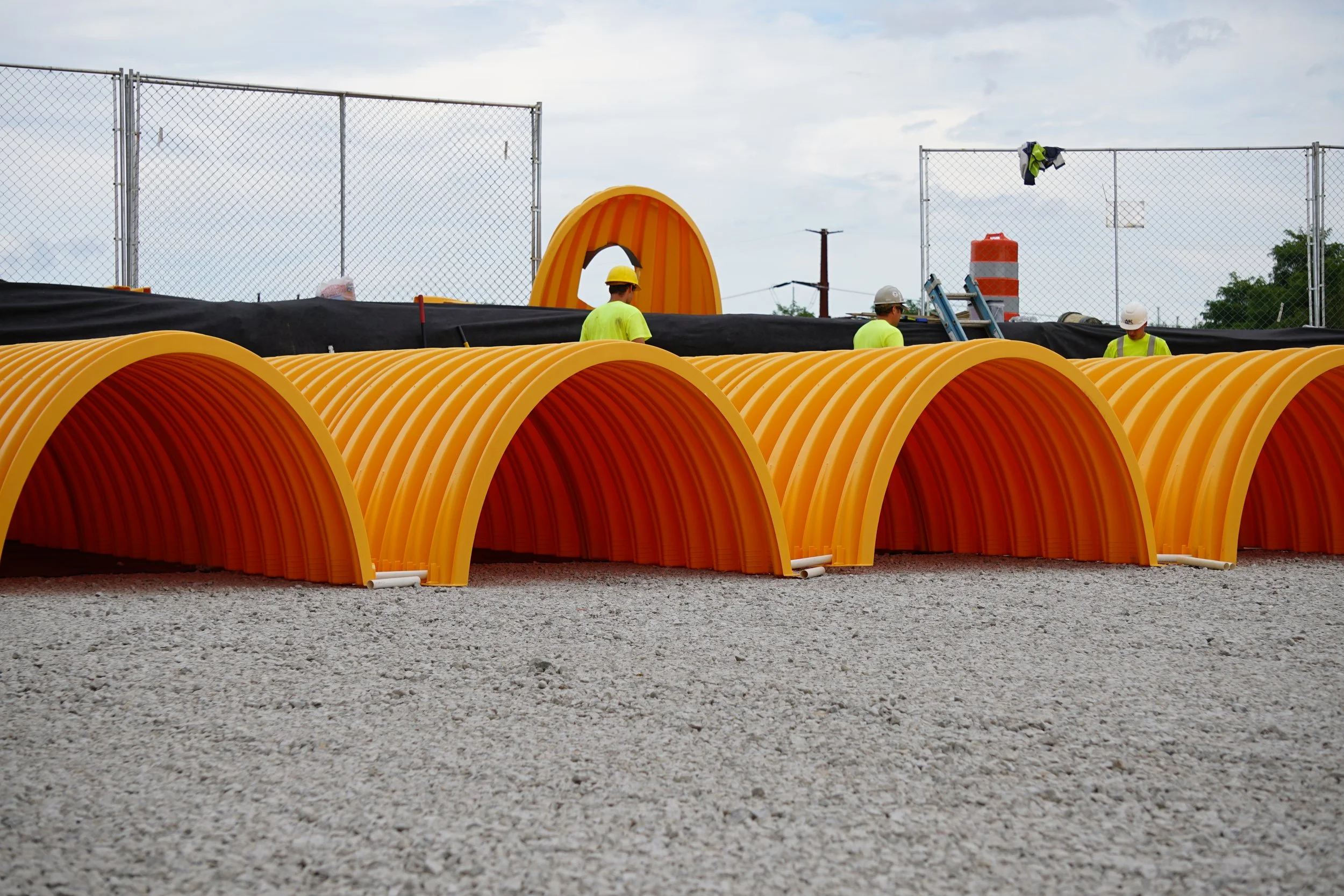 Construction site with orange curved pipes, workers in yellow and white helmets, black sheet covering pipes, temporary fencing, orange barrel, ladder, and cloudy sky.