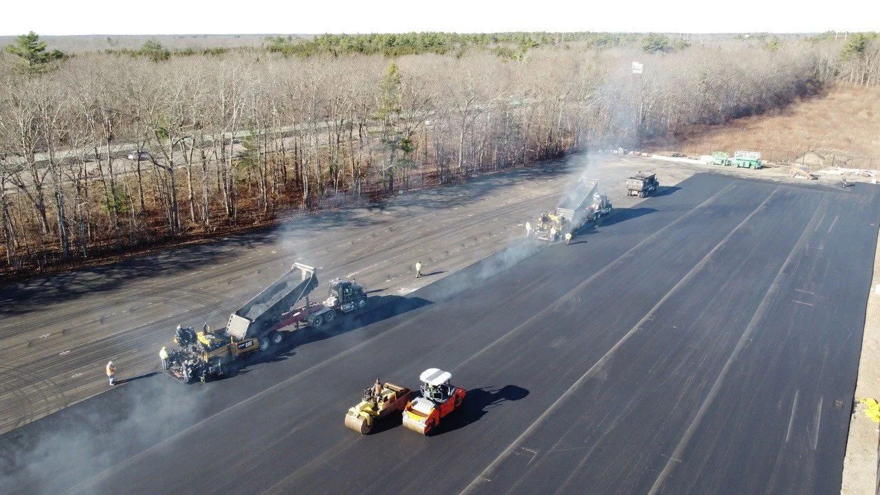 Extensive highway paving operation with asphalt rollers, dump trucks, and road construction workers laying new asphalt on a large highway, surrounded by trees and open land.