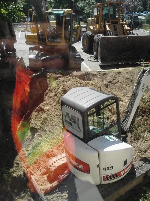 Construction site with a small Bobcat mini excavator digging in the dirt, and two larger yellow bulldozers parked behind it.