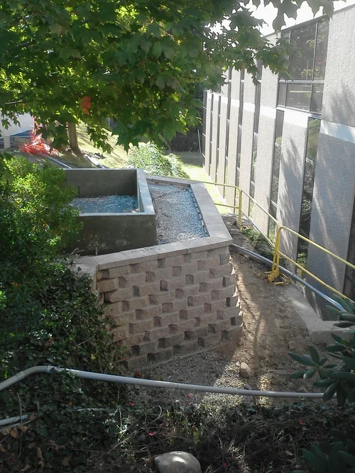 Construction site with concrete water feature and brick foundation, yellow safety railing, nearby trees and a building with siding.
