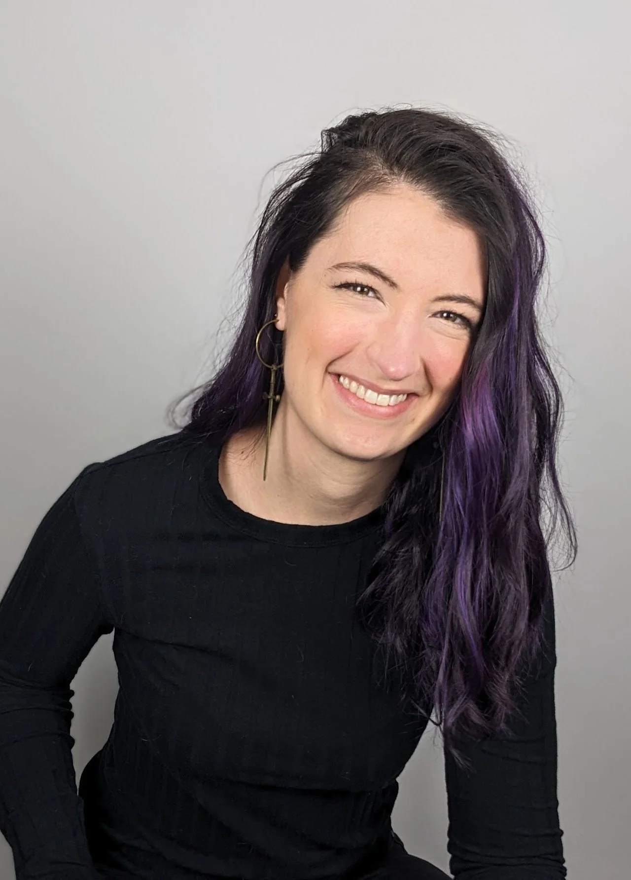A woman with long dark hair with purple highlights, wearing a black top and gold earrings, smiling against a plain light gray background.