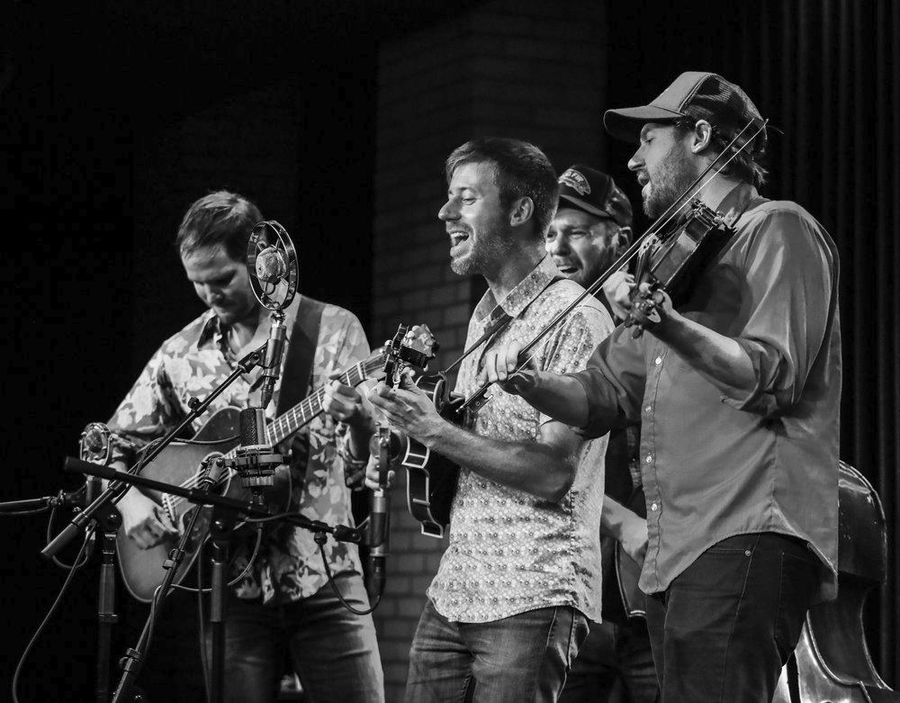 Group of four men performing music on stage with guitars and a microphone, one playing fiddle, in black and white.