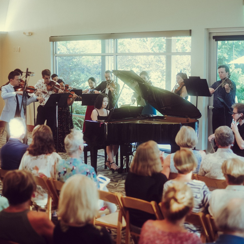A classical music concert with a pianist playing on a grand piano and a string ensemble including violinists and a cellist performing in front of an audience in a bright room with large windows.