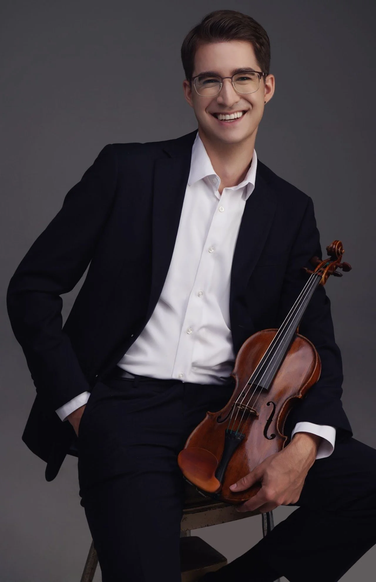 A young man in a black suit and white shirt sitting and holding a violin, smiling, against a dark gray background.