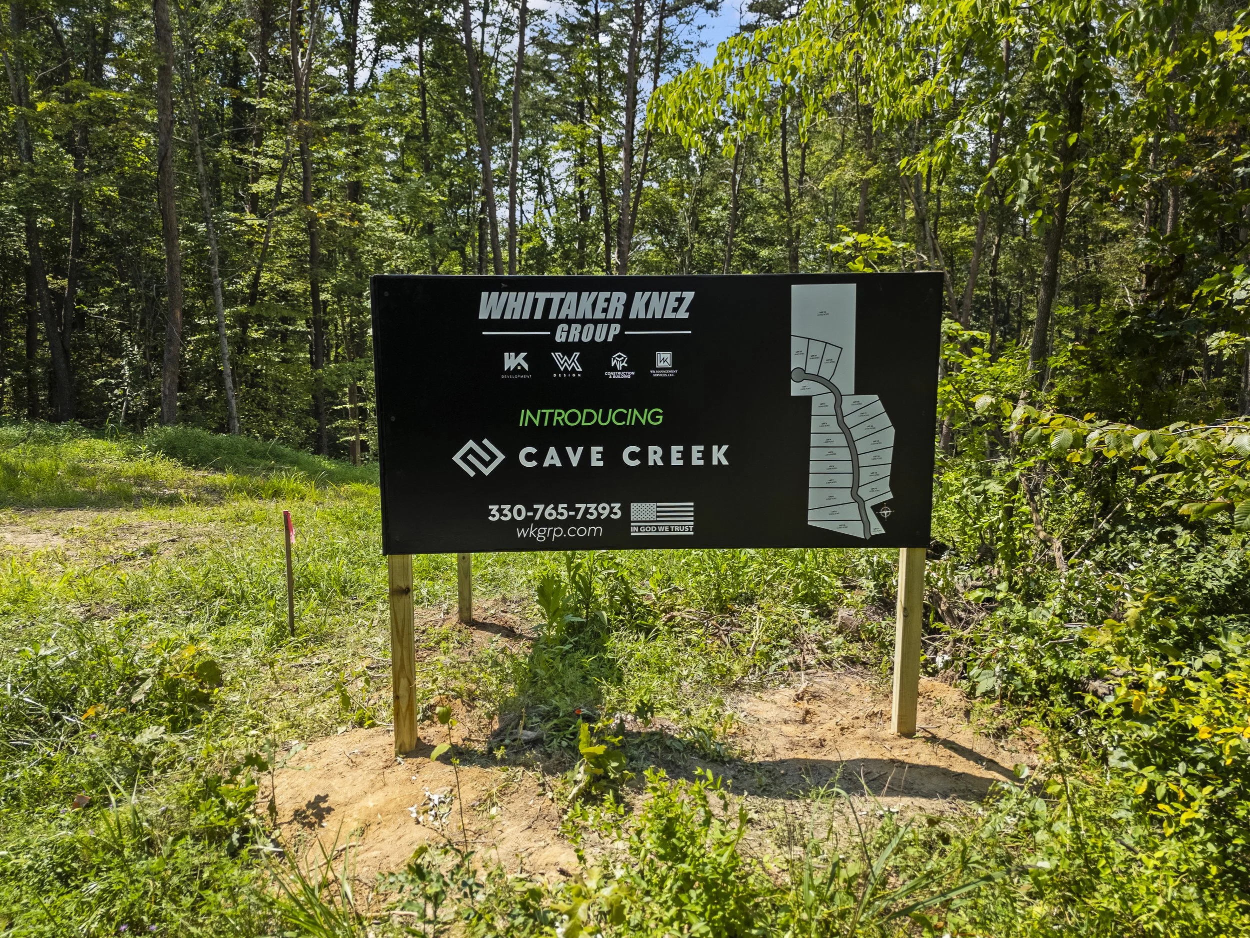 A large black sign with white and green text announcing the introduction of Cave Creek by Whittaker Knez Group, with a map on the right, contact information, and logo icons, placed on wooden posts in a grassy area with trees in the background.