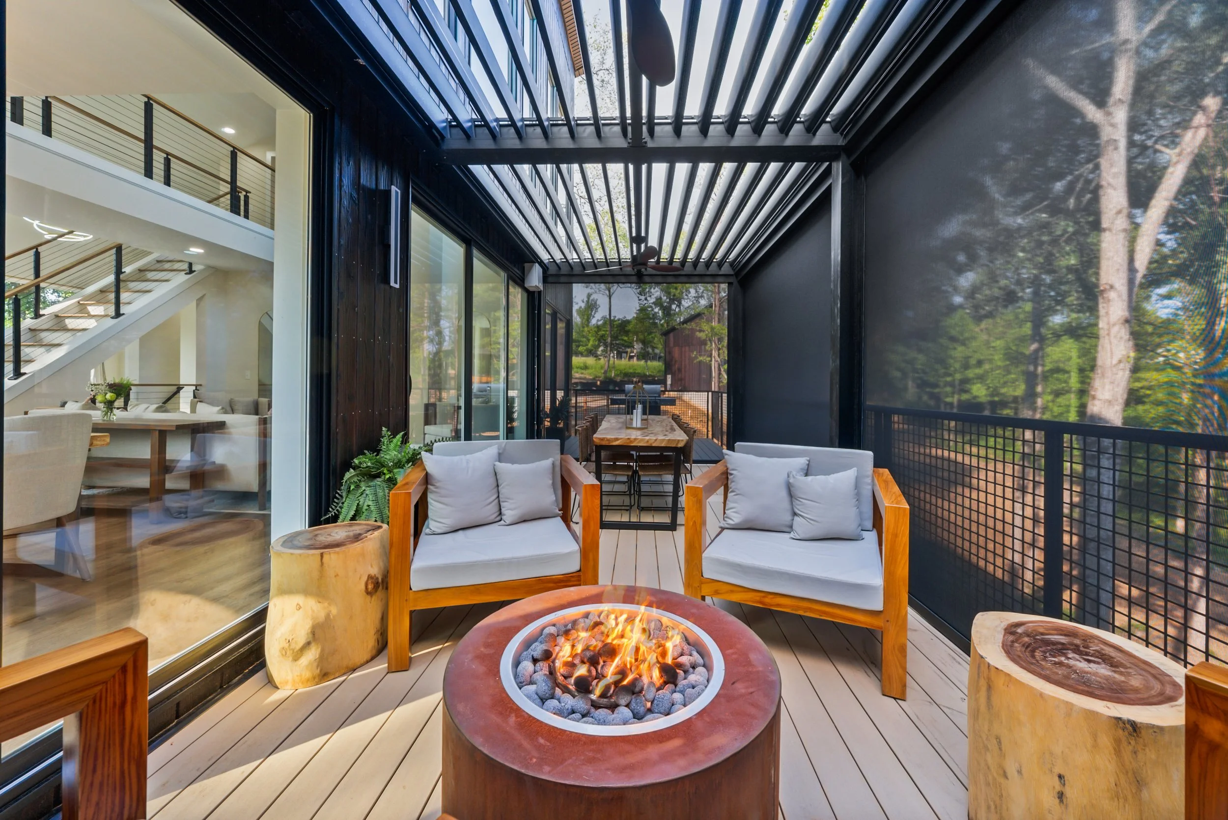Outdoor balcony with two wooden-framed chairs with white cushions, a fire pit with flames, and two wooden tree stump side tables, overlooking a landscaped yard with trees.