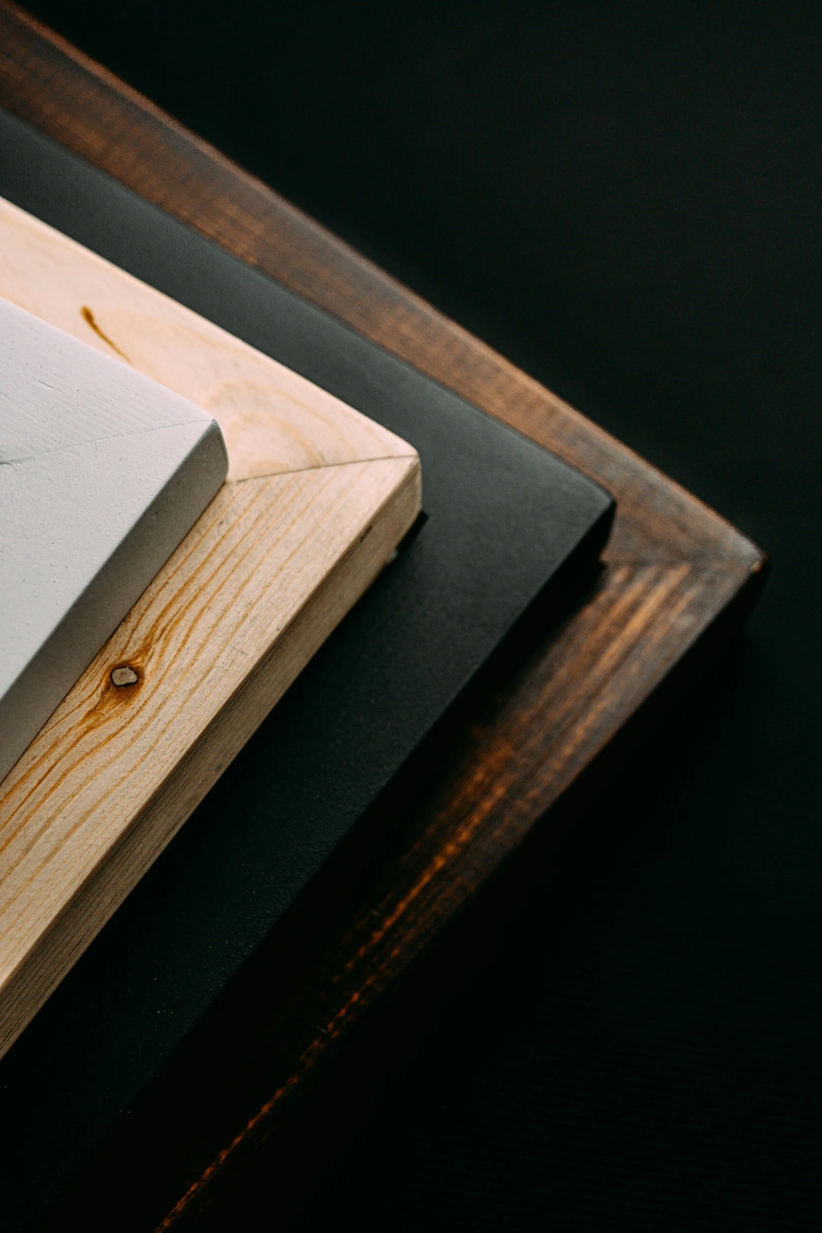 Close-up of stacked picture frames in different colors and finishes, including white, light wood, black, and dark wood, on a dark surface.