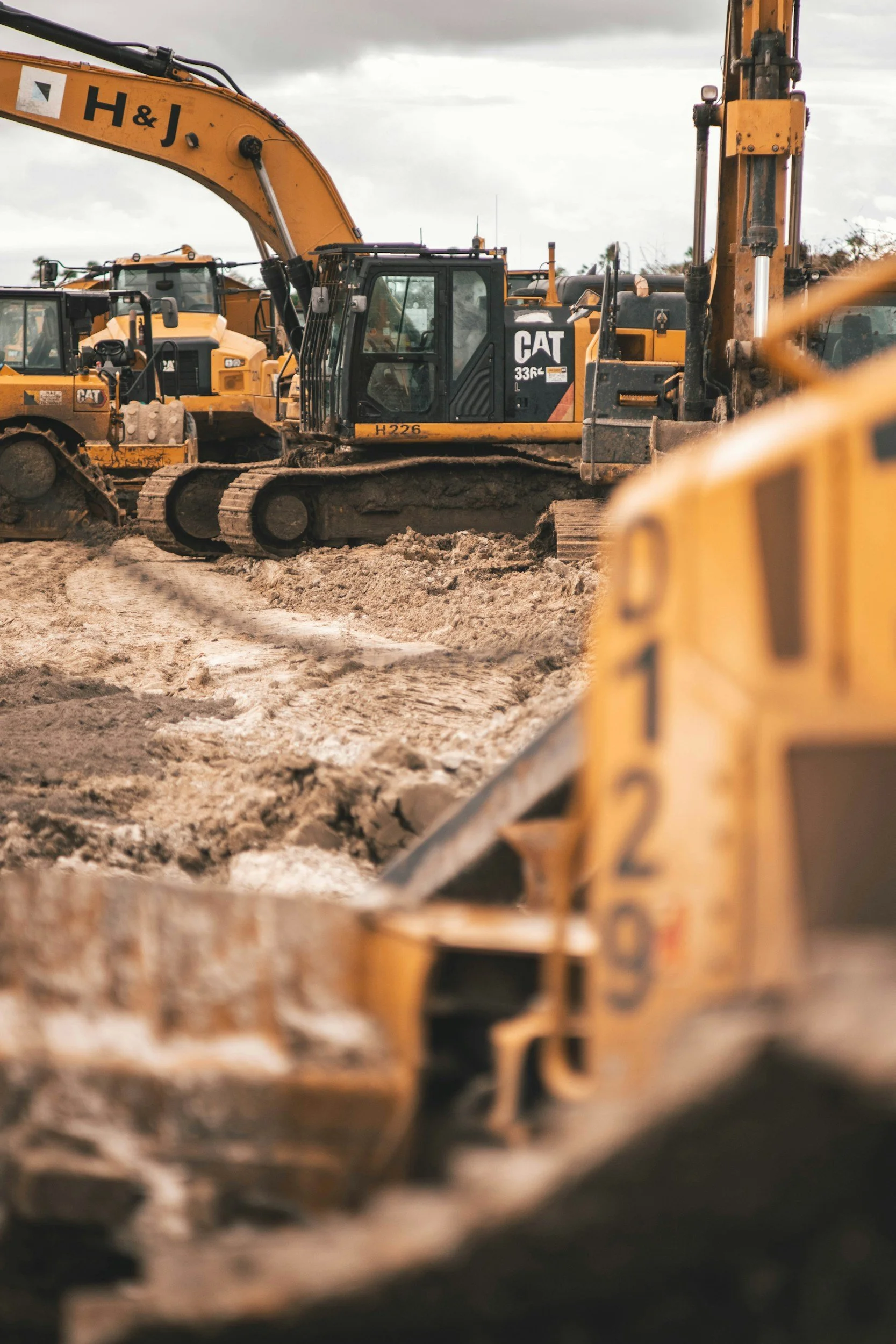 Construction site with multiple yellow excavators, one prominently labeled 'CAT 336E' and 'H&J', working on rough, dirt terrain under a cloudy sky.