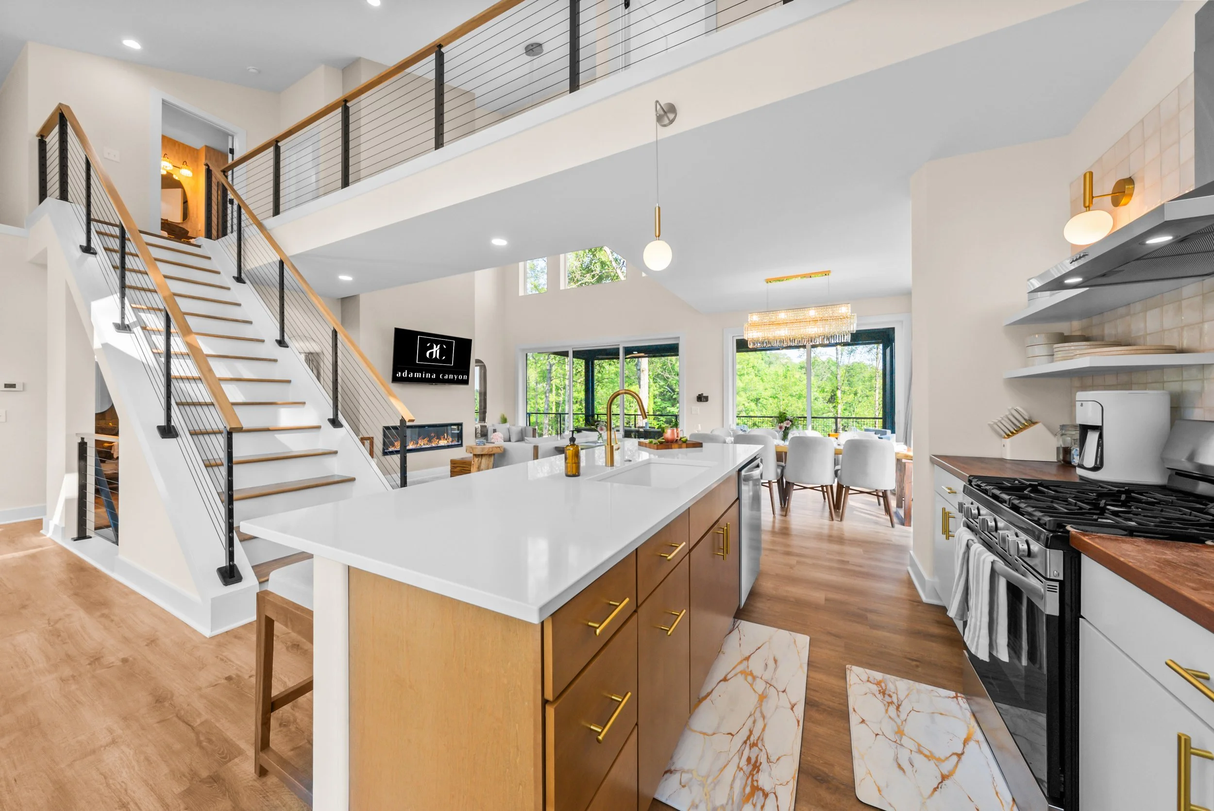 Open-concept kitchen with a large white island, wooden cabinets, stainless steel appliances, and hardwood floors. Bright natural light from windows, modern lighting fixtures, and a view of greenery outside.