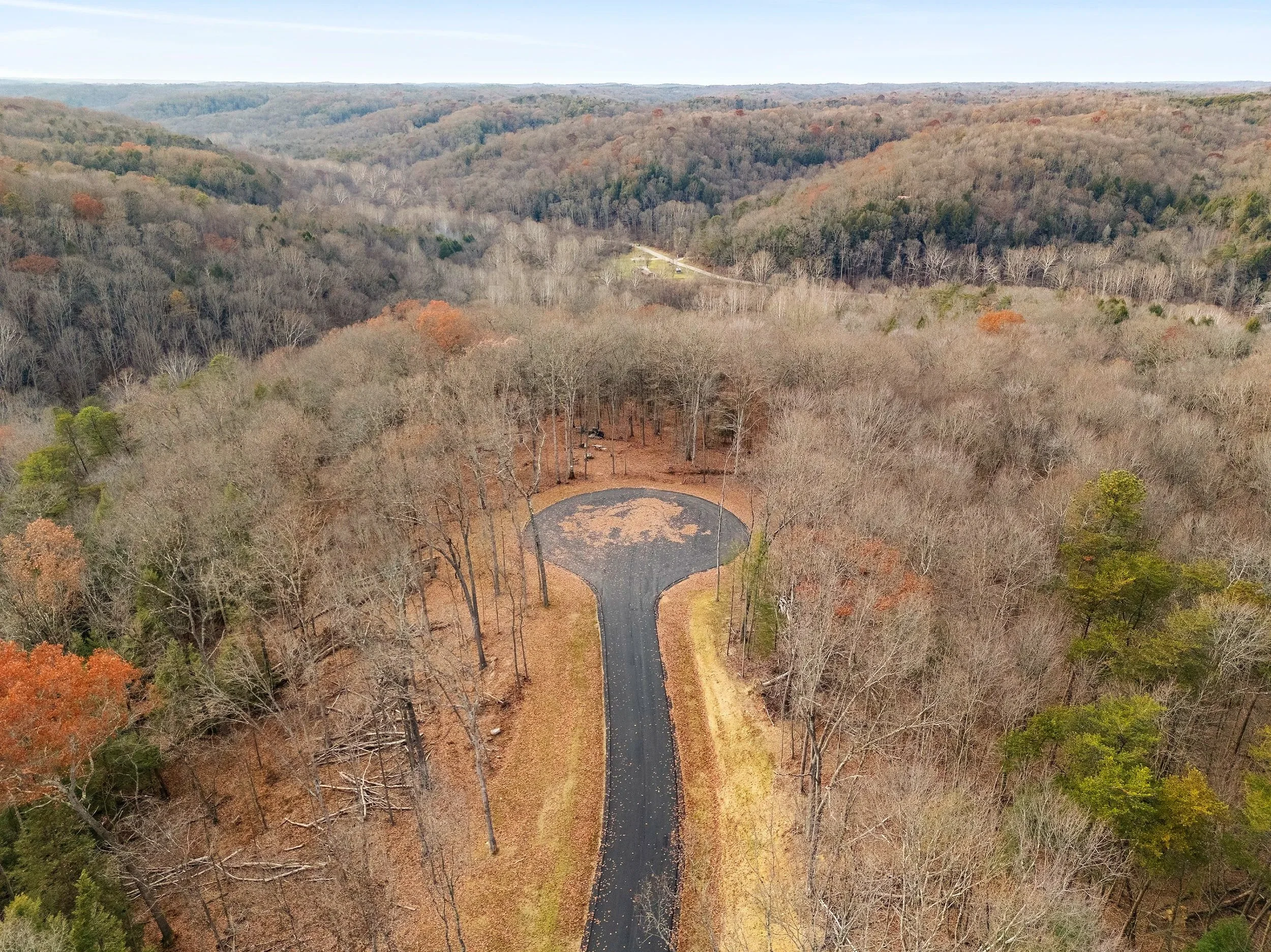 An aerial view of a winding paved road leading to a circular clearing surrounded by leafless trees in a forested mountainous area during fall.