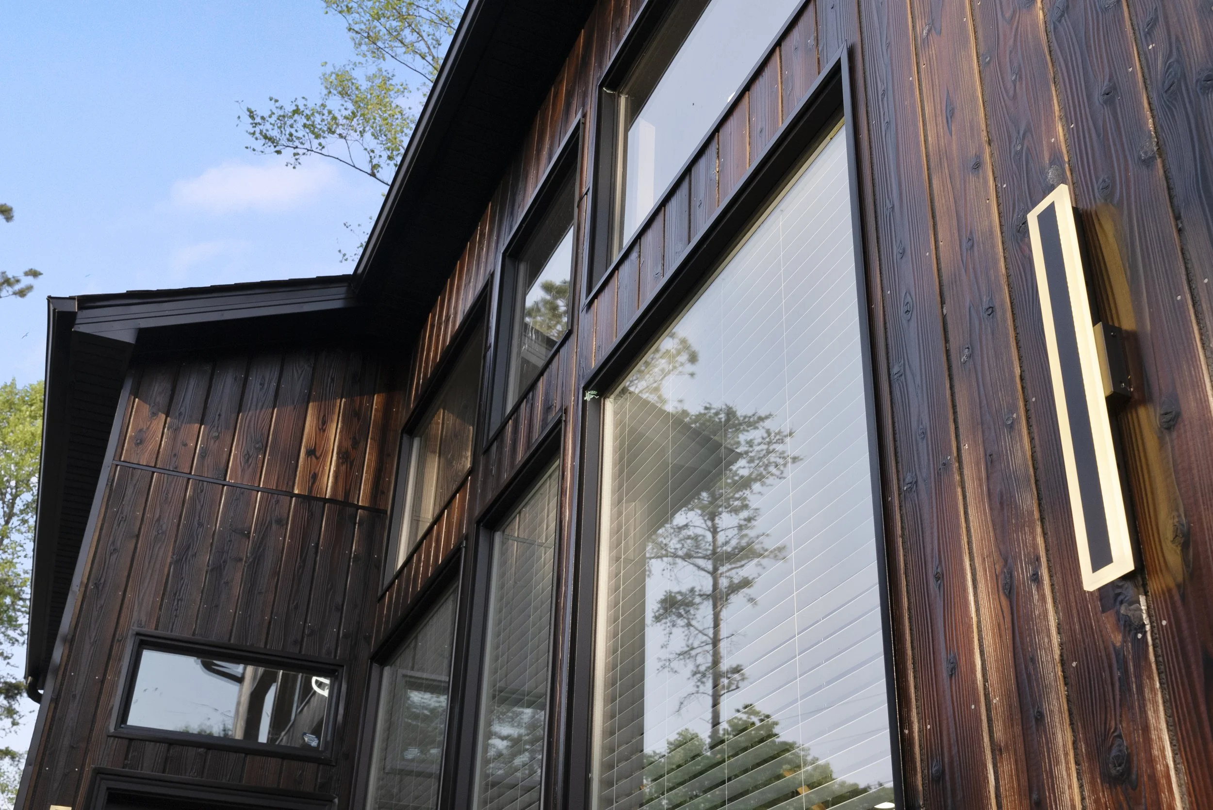 Close-up of a modern house with a dark wood exterior and large windows reflecting trees and sky, under a blue sky with some clouds.