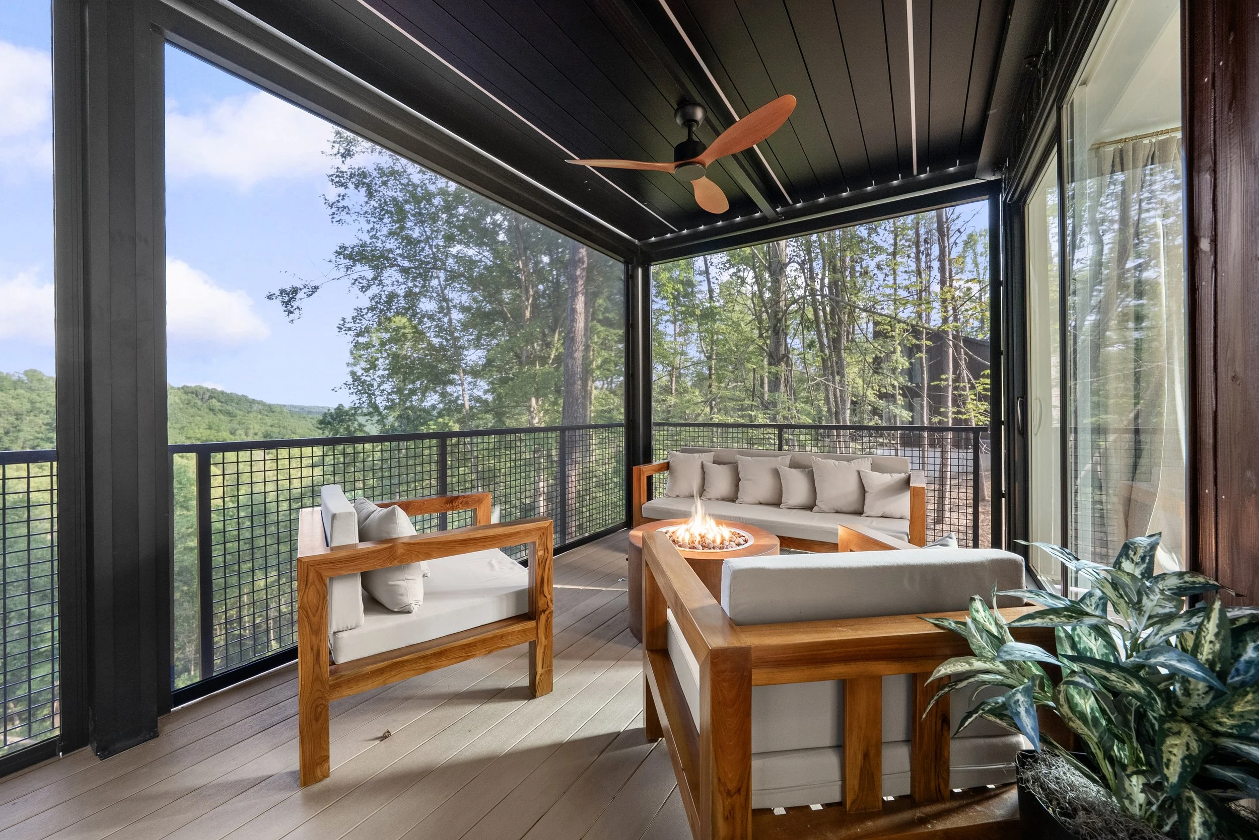 screened porch with wooden furniture, beige cushions, a fire pit, ceiling fan, and a view of trees and woods outside.