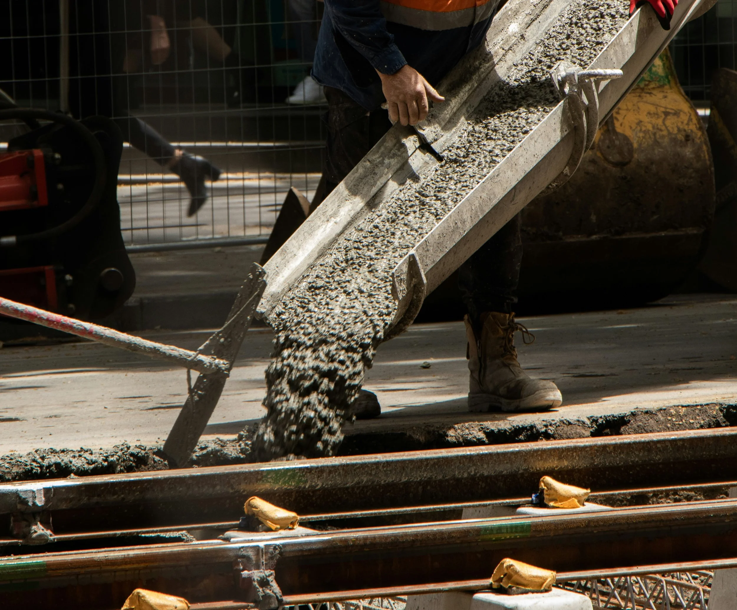Worker pouring concrete onto a construction site using a chute. The worker is wearing brown boots and a dark blue jacket, with other construction materials and equipment visible in the background.
