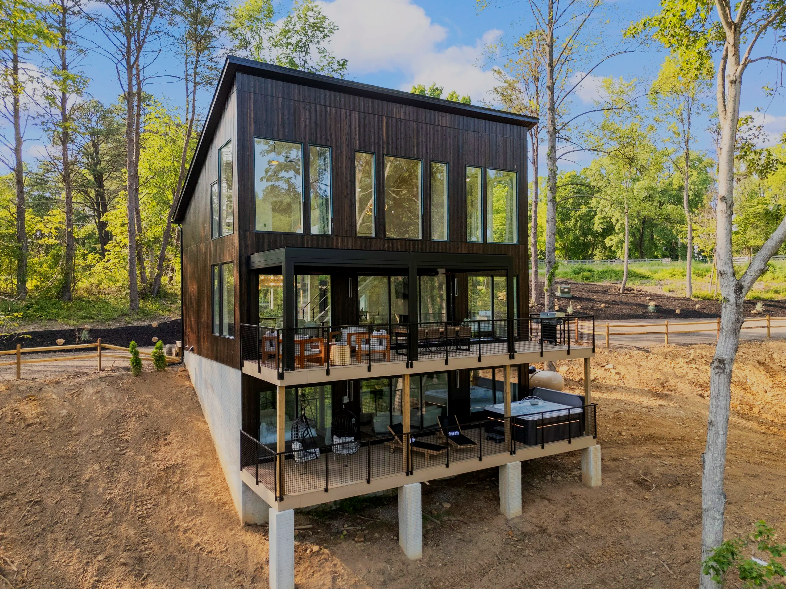 Modern, multi-story house with black exterior and large windows, built on stilts on a sloped terrain surrounded by trees.