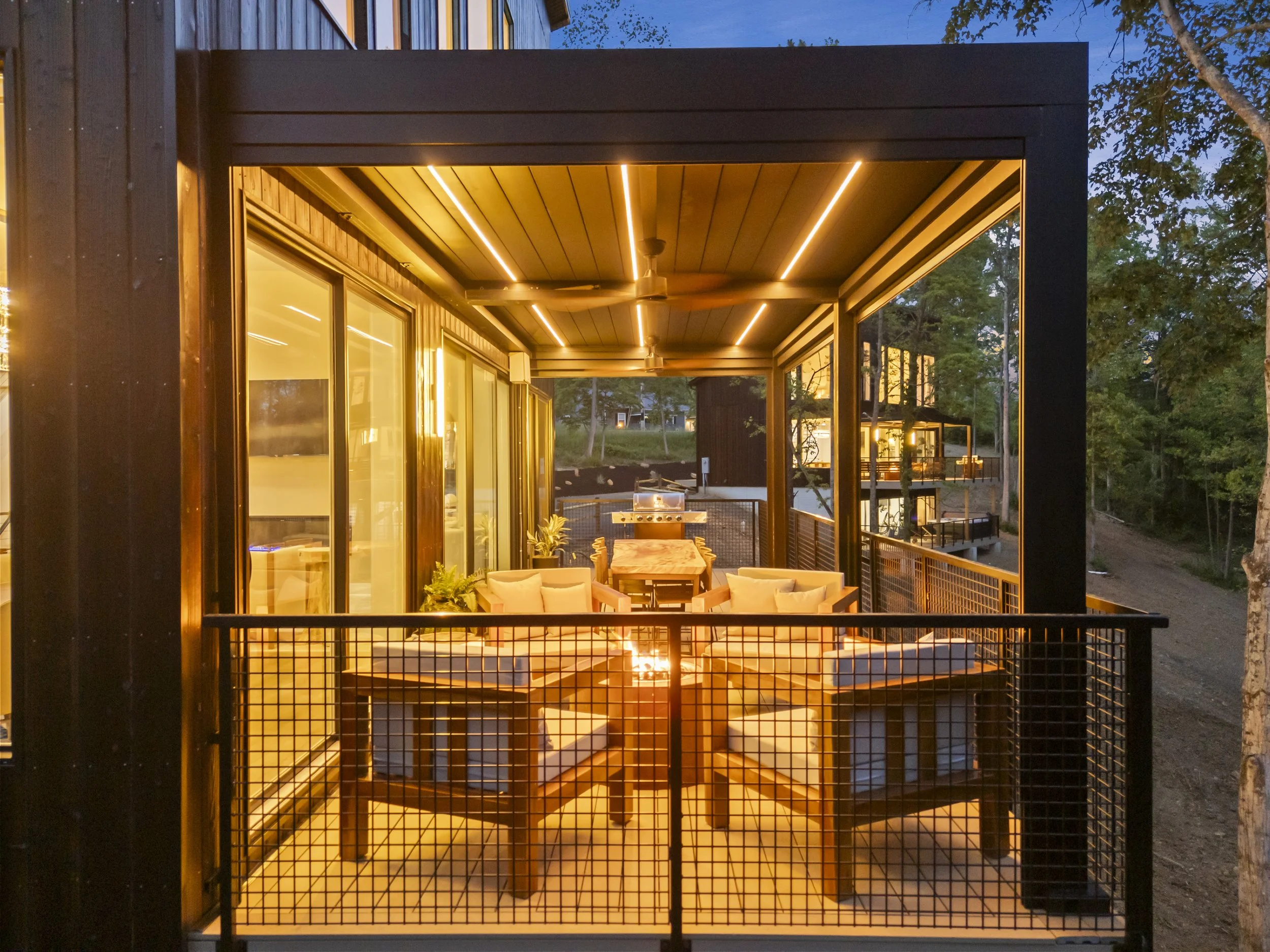 Outdoor patio at dusk with seating around a fire pit, glass sliding doors, and modern house exterior with black metal framing and wood accents, overlooking a wooded area.