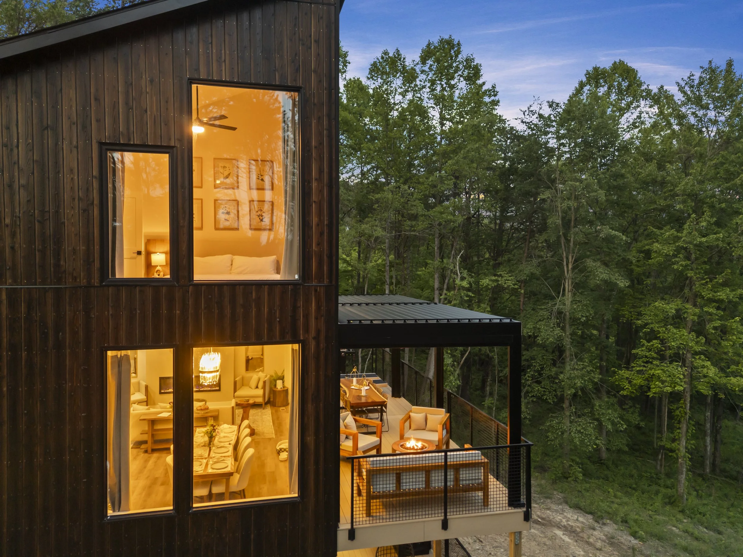 A modern house with dark wooden exterior walls, large glass windows revealing a cozy interior with warm lighting, a dining area, living room, and a balcony with outdoor furniture and a fire pit, surrounded by lush green trees during dusk.