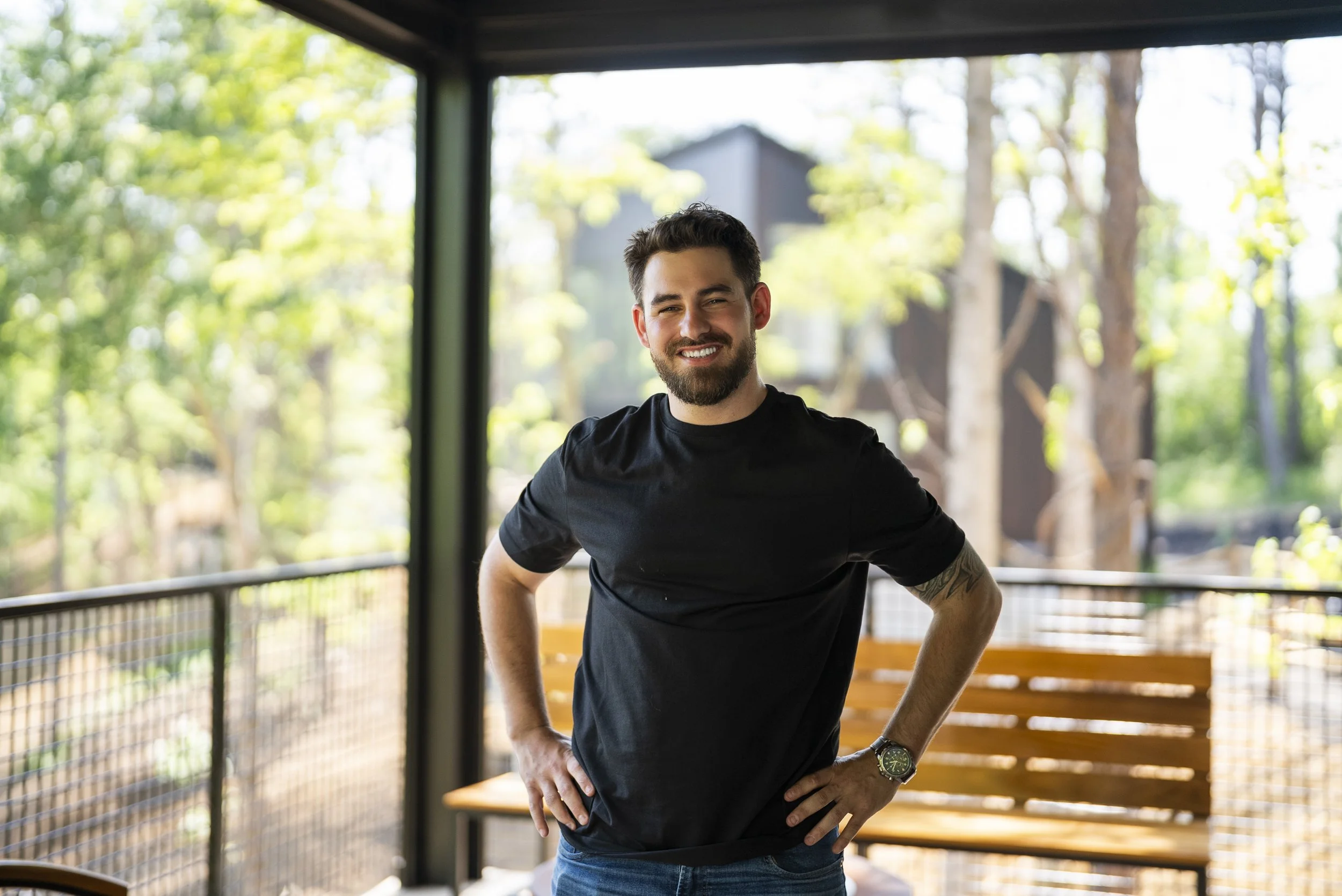 Smiling man with dark hair, beard, and mustache wearing a black t-shirt and jeans, standing outdoors on a wooden deck with trees and houses in the background.