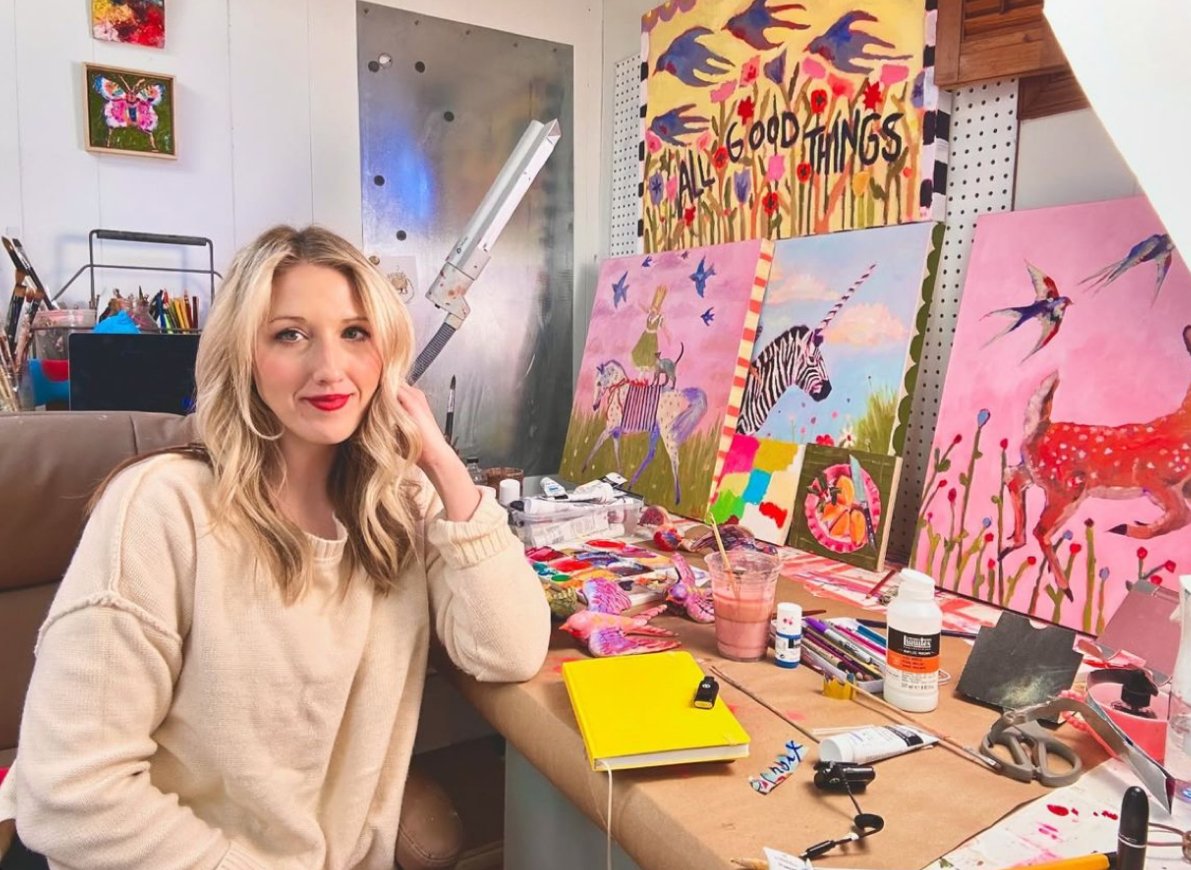 A woman with blond hair and red lipstick sitting at an art studio desk with colorful paintings of animals and playful scenes, including a zebra, flamingos, and a deer. The workspace has paint supplies, a pink beverage, a yellow notebook, and art tools, with a large colorful painting in the background that says "All Good Things."