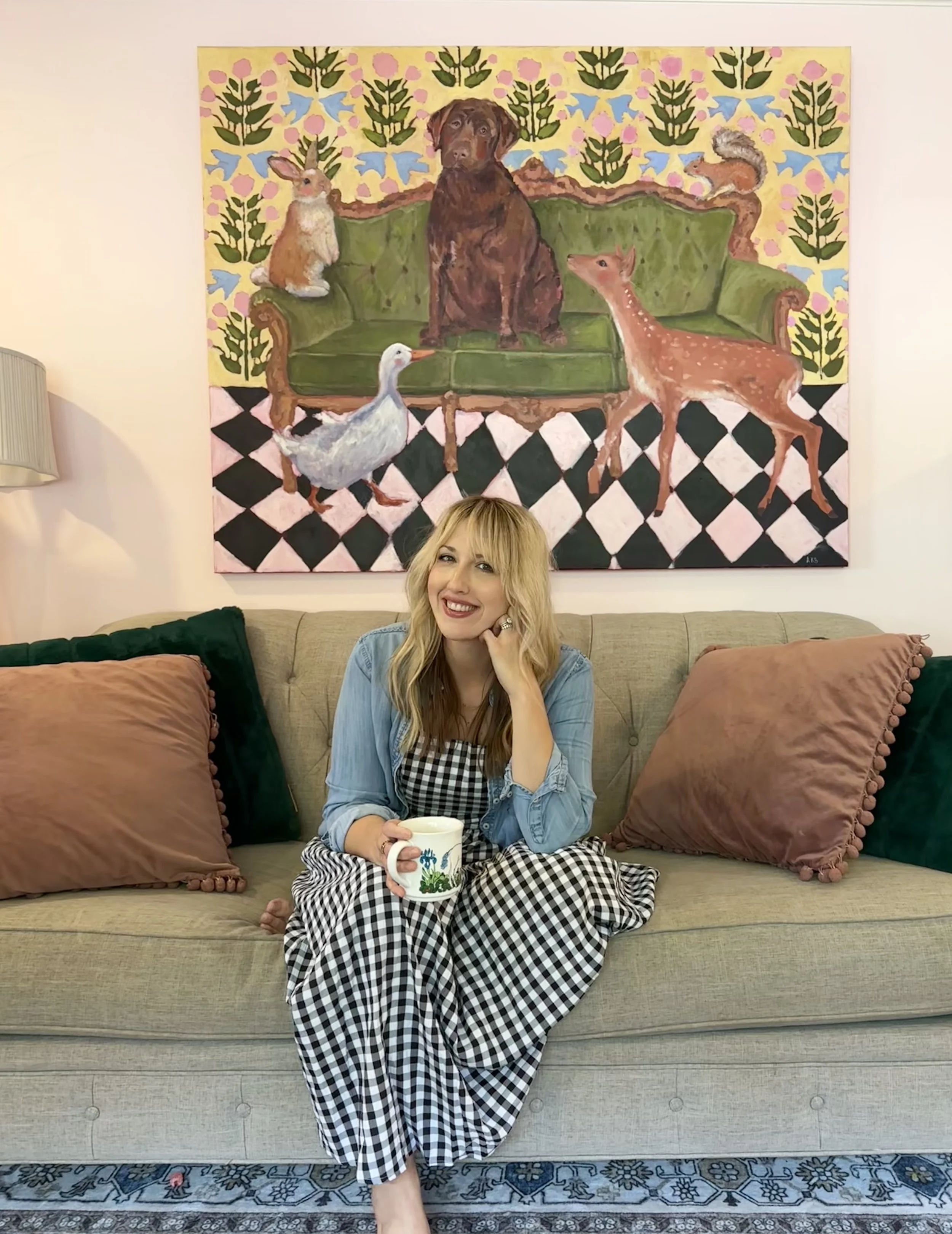 A woman with blonde hair in a gingham dress and denim jacket smiling, sitting on a beige sofa with pink and dark green pillows, holding a mug with a floral design, in a living room with a large colorful painting of animals hanging on the wall behind her.
