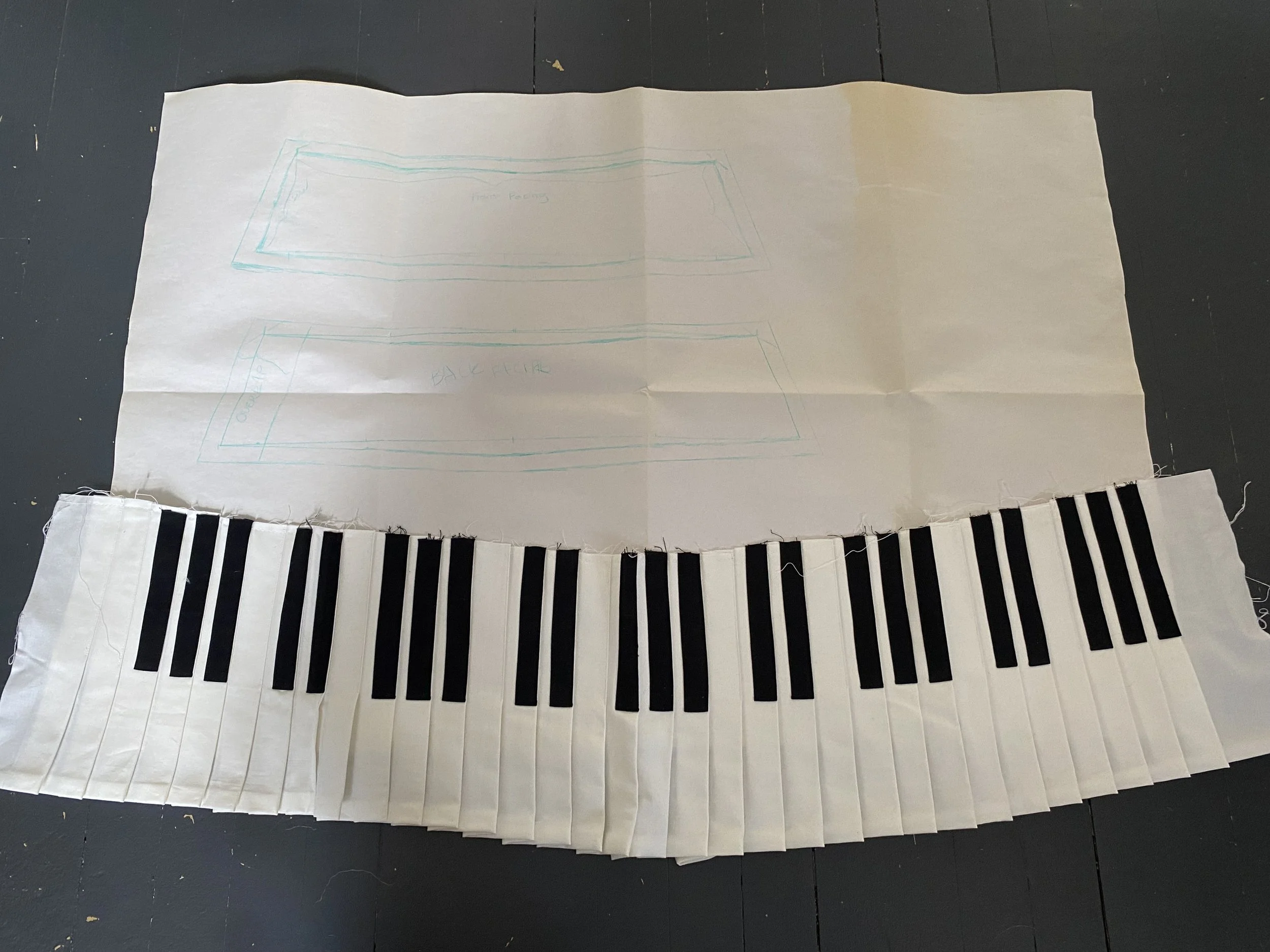A large sheet of paper with a hand-drawn sketch of two rectangles labeled 'the facing' and 'back face.' There is a fabric cutout of a piano keyboard with black and white keys attached to the bottom edge of the paper, on a dark surface.