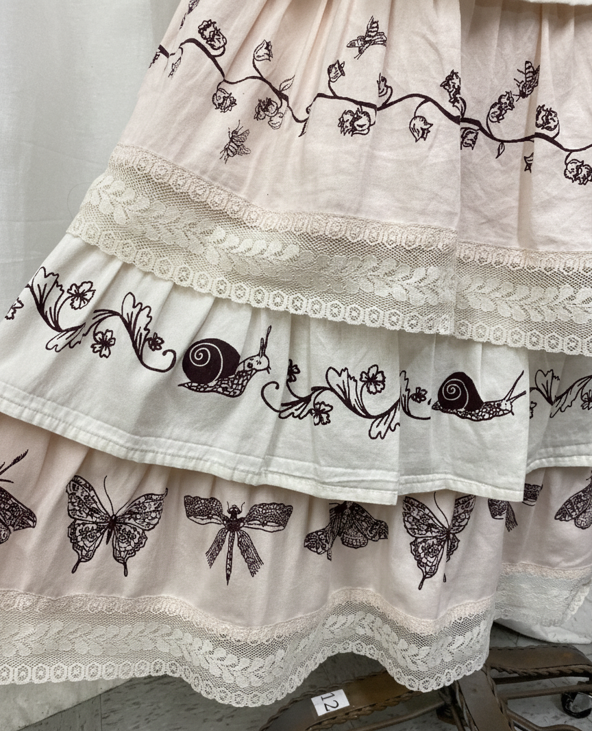 A white dress with black embroidered designs, including snails, butterflies, flowers, and insects. The dress features lace and decorative trim details.