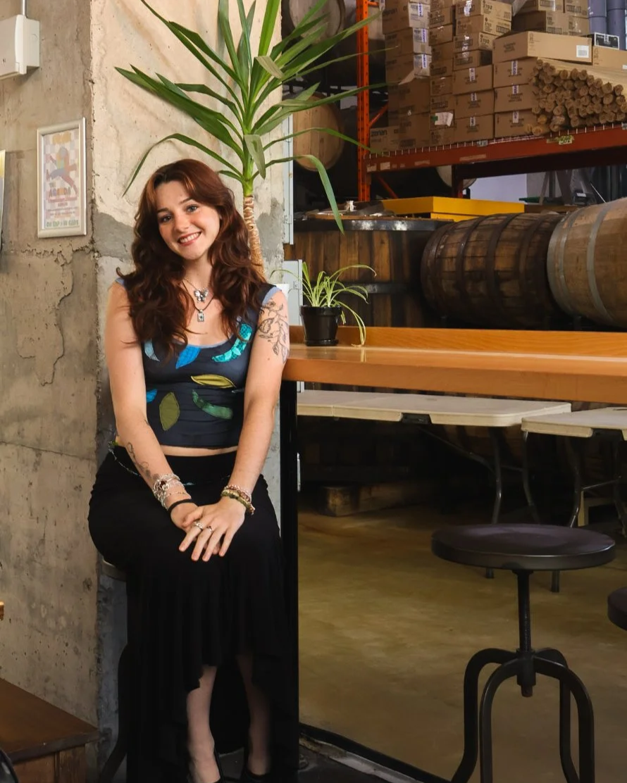 A young woman with curly red hair and tattoos on her arms smiling and sitting on a stool in a rustic, industrial-style space with wooden barrels and shelves stocked with boxes in the background.