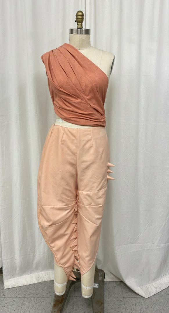 A mannequin wearing a one-shoulder rust-colored top with pleats and sash detail, paired with oversized blush pink pants with ruched detailing and spike embellishments on the side.