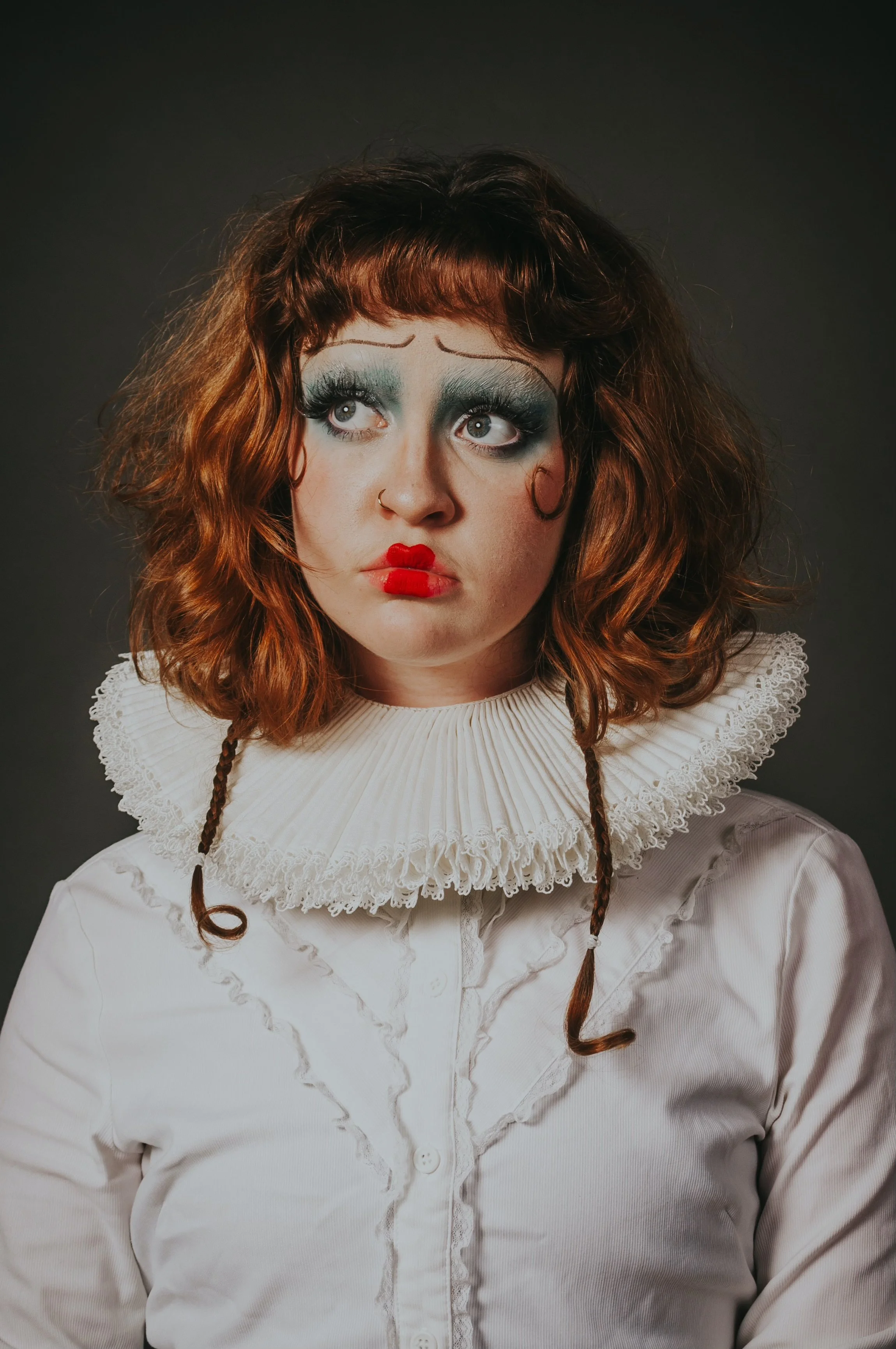 A woman with curly red hair, exaggerated makeup, wearing a puffy white ruffled collar and a white blouse, looks upward with a concerned expression against a dark background.
