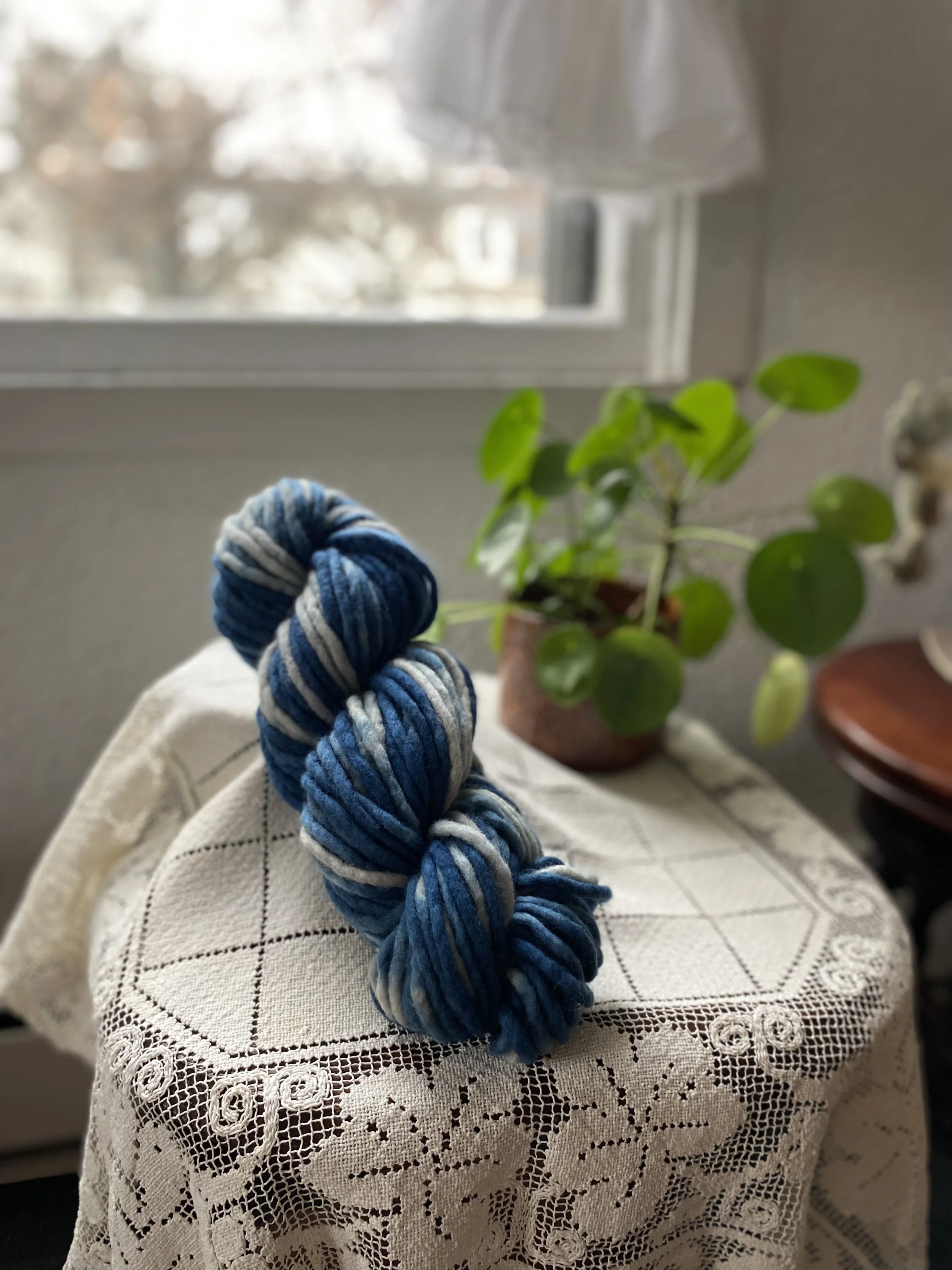 A skein of blue and gray yarn resting on a lace-covered table next to a potted plant with round green leaves, with a window in the background.