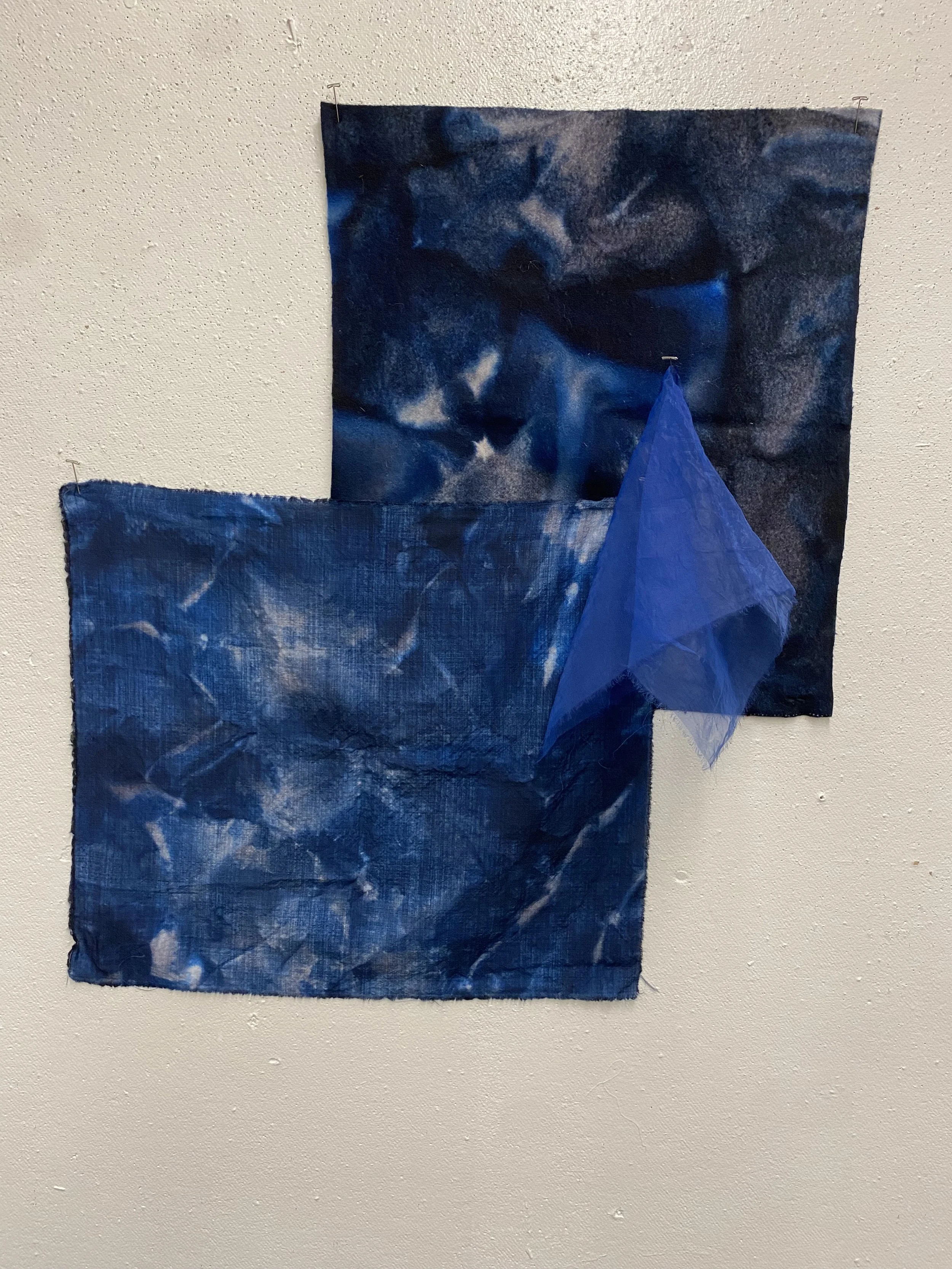 Two square fabric pieces and a blue cloth hanging on a white wall.