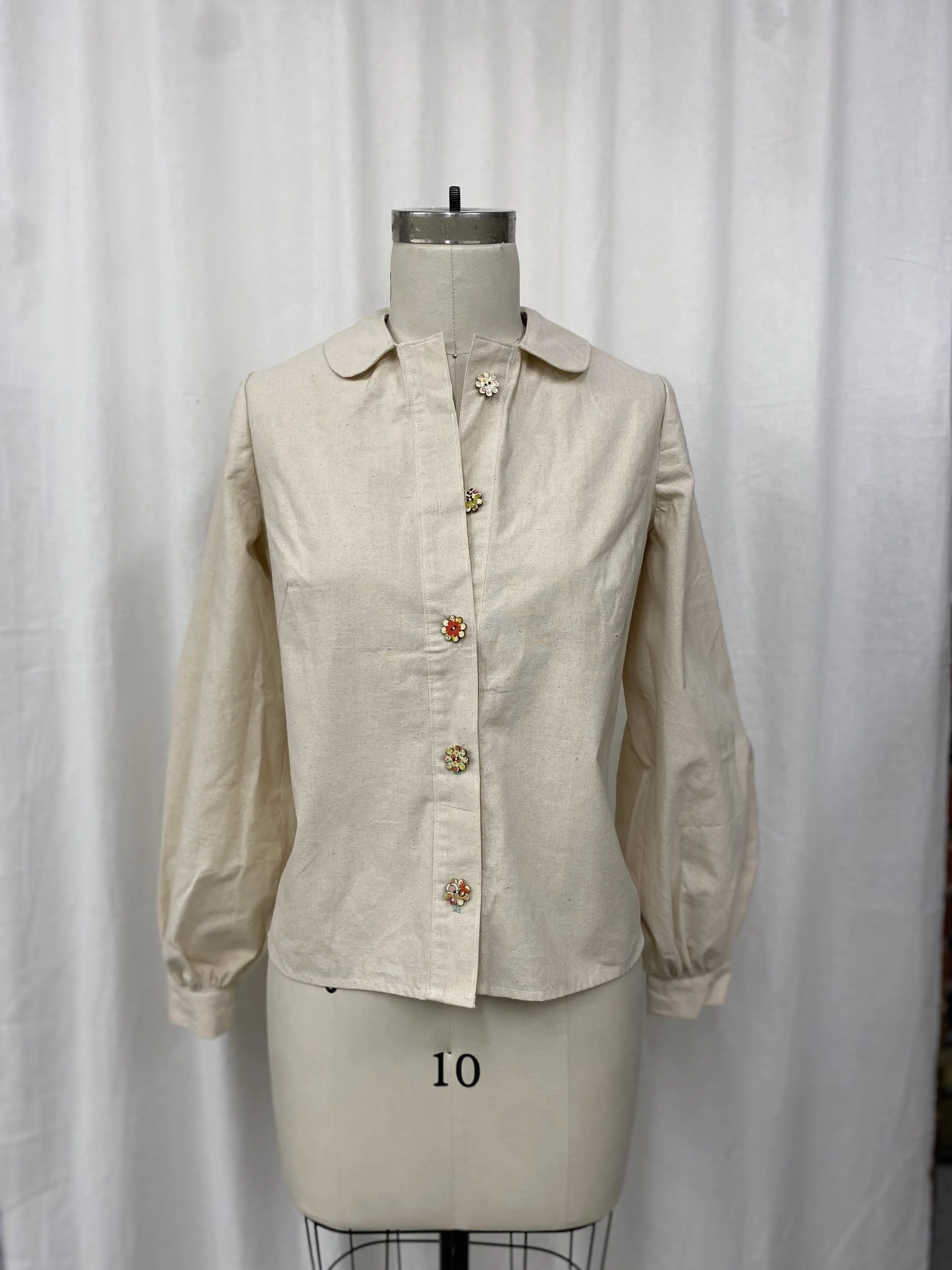 Beige button-up shirt with floral embellishments on the buttons, displayed on a dress form stand in front of a white curtain.