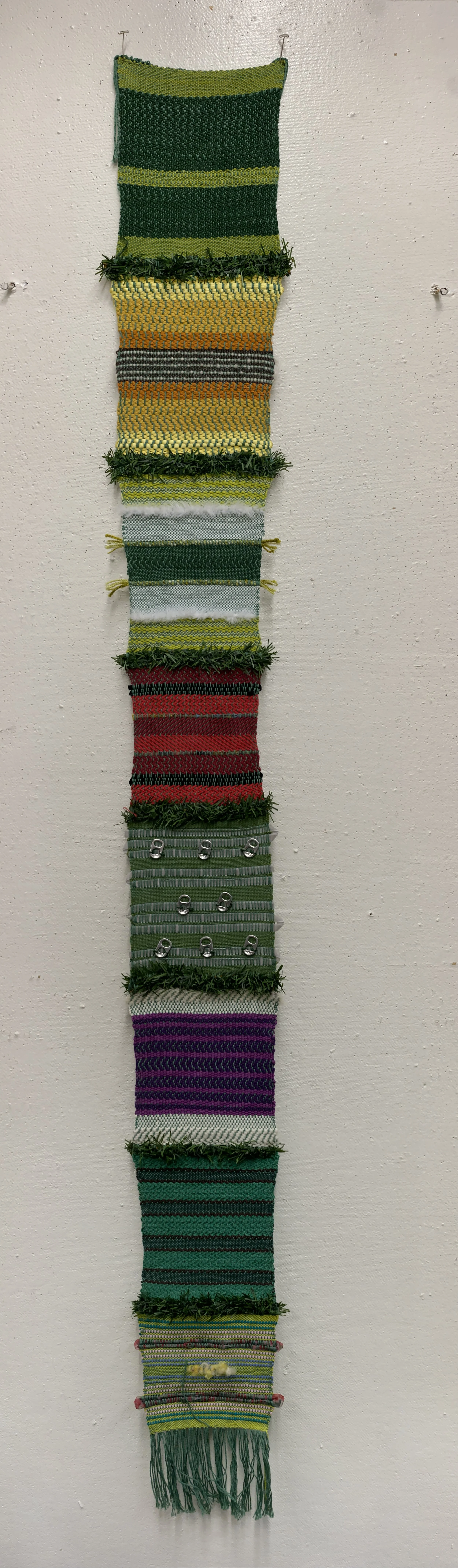 Vertical display of colorful woven fabric strips on a wall, featuring green, yellow, orange, red, purple, and gray colors, some with fringes, attached with small hooks.