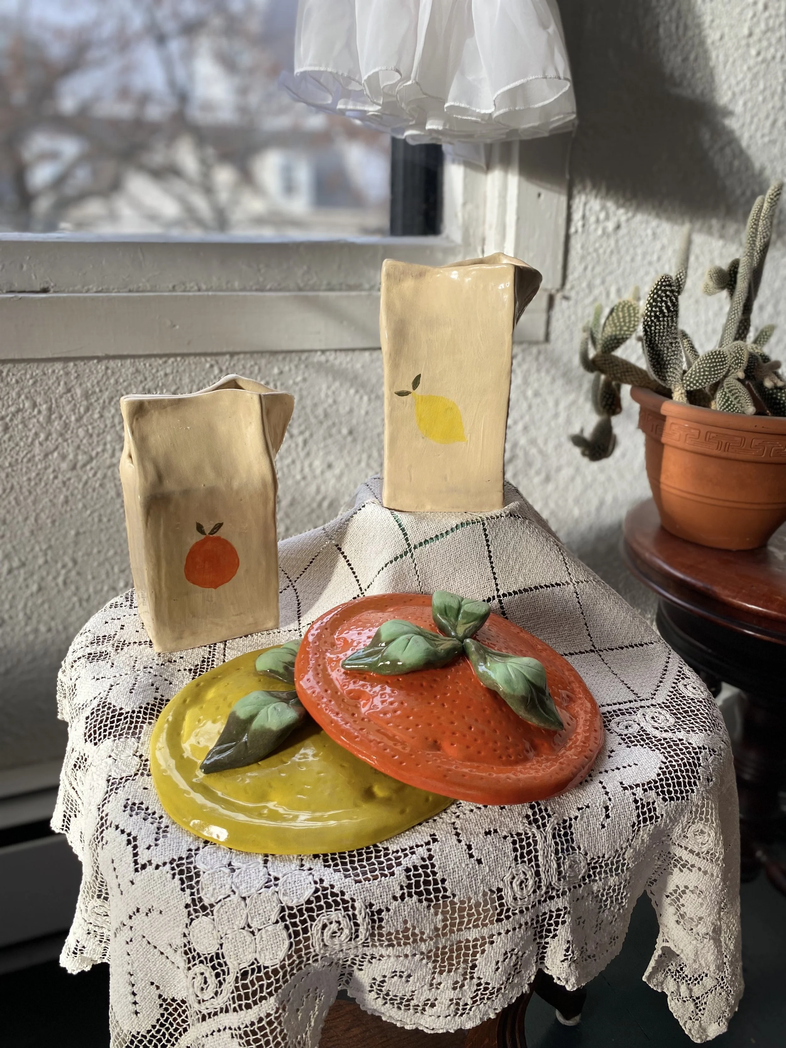 Decorative ceramic fruits and two painted ceramic cartons on a lace-covered table next to a potted cactus plant near a window.