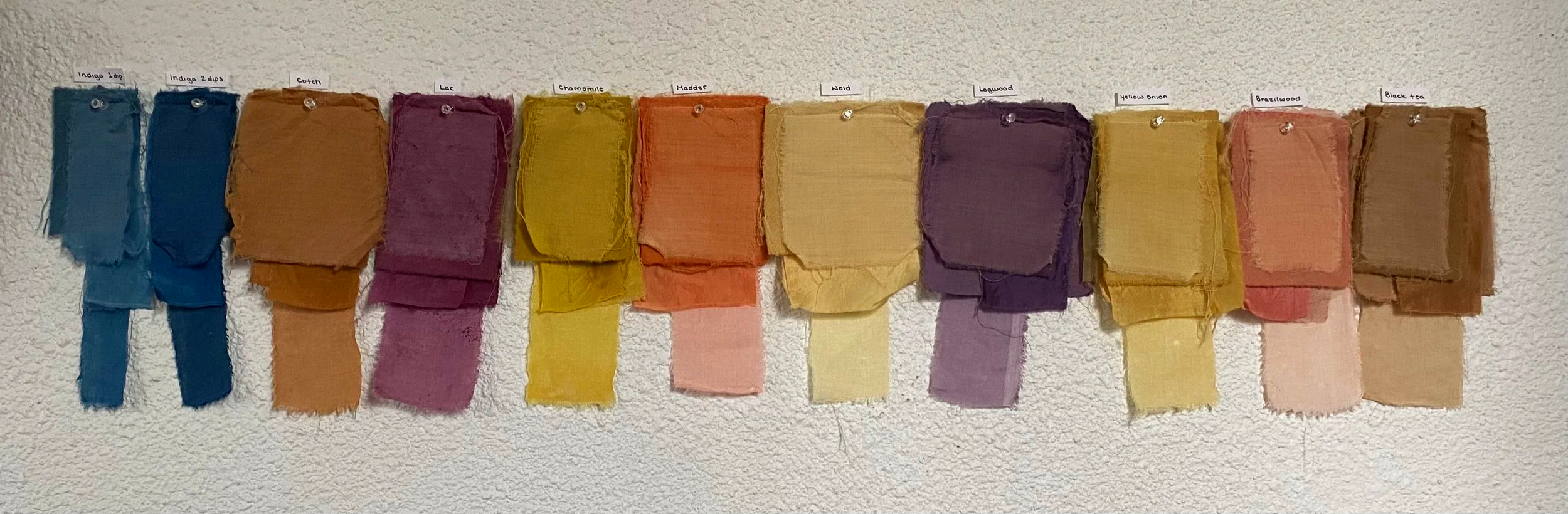 Colorful fabric swatches hanging on a wall, labeled with color names such as Indigo 1ap, Cutch, Lac, Chamomile, Madder, Weld, Logwood, Yellow Onion, Brazilwood, and Black Tree.