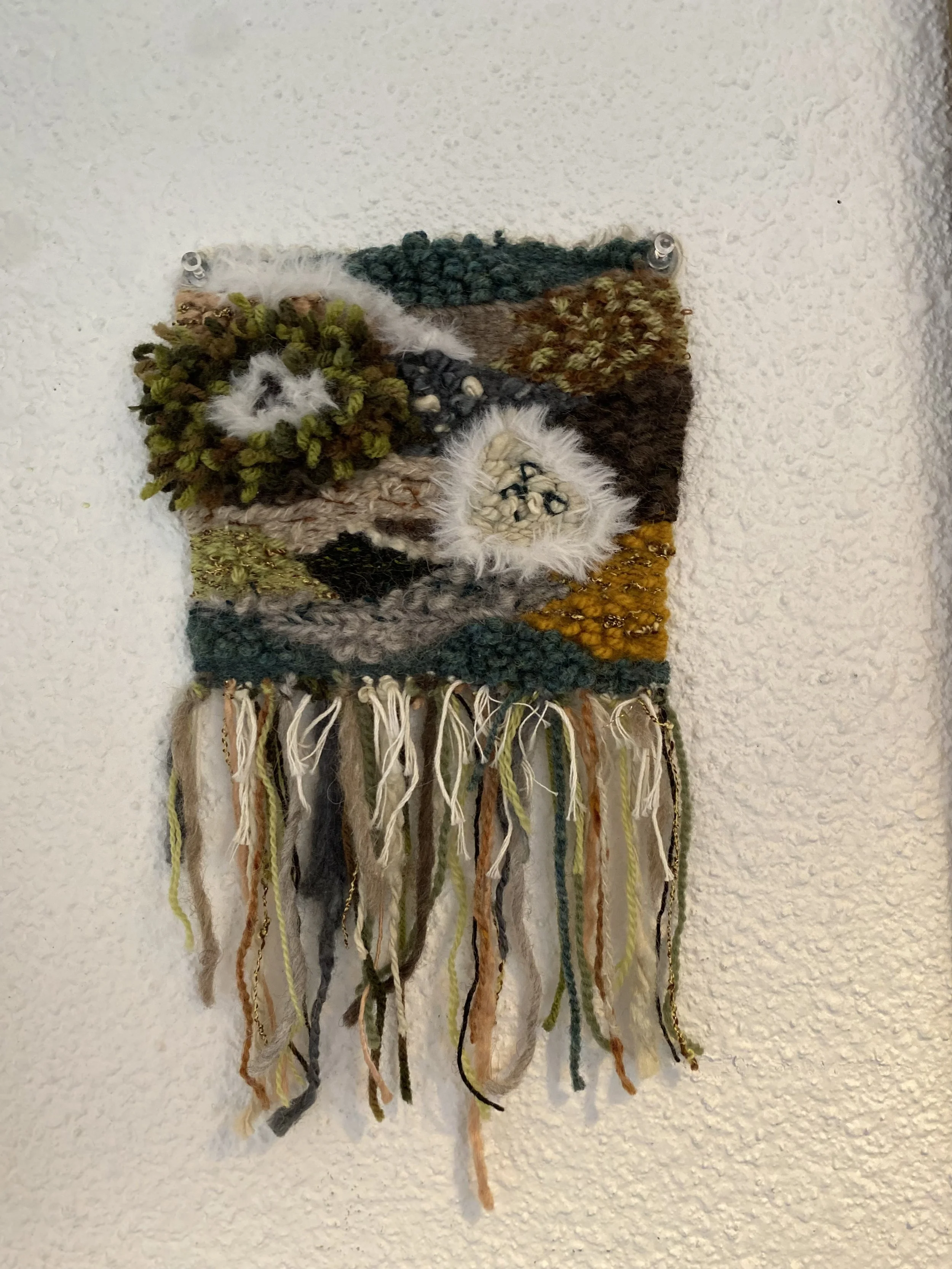 Colorful woven textile wall hanging with textured yarns, feathers, and fringe on a white textured wall.
