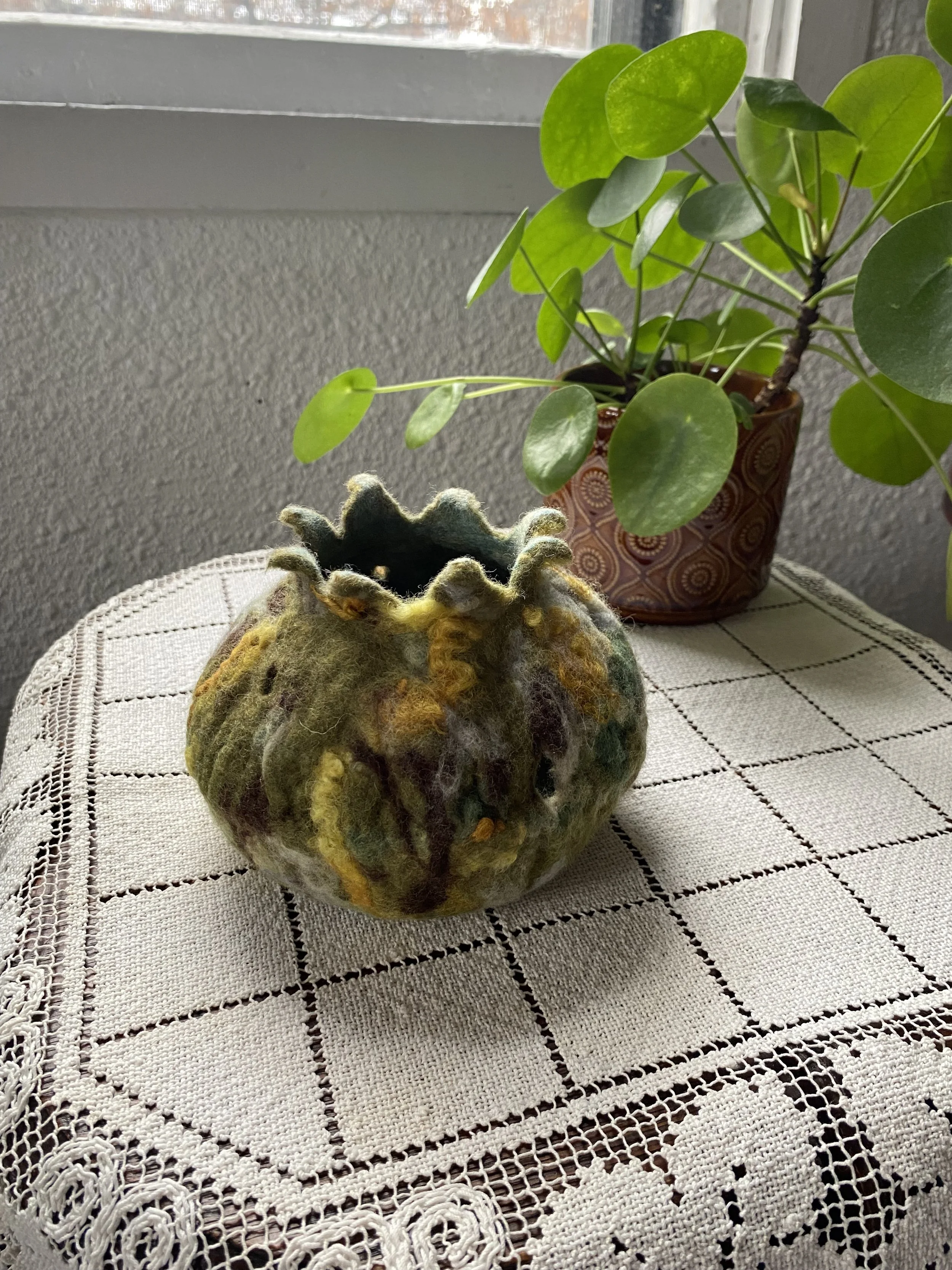 A felted yarn pumpkin with green, yellow, and brown colors sitting on a crocheted tablecloth, with a potted plant with round green leaves in the background near a window.