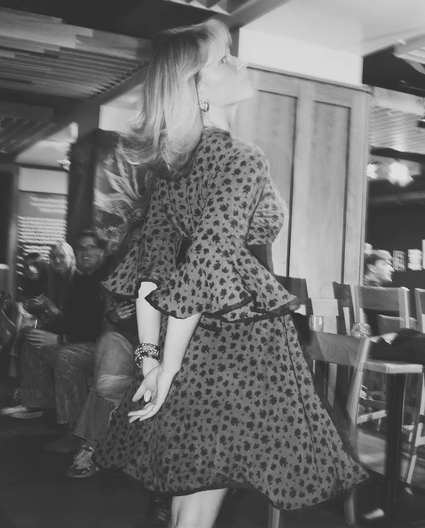 A woman in a leopard print dress with ruffled sleeves standing in a restaurant or cafe, with a few people seated in the background.