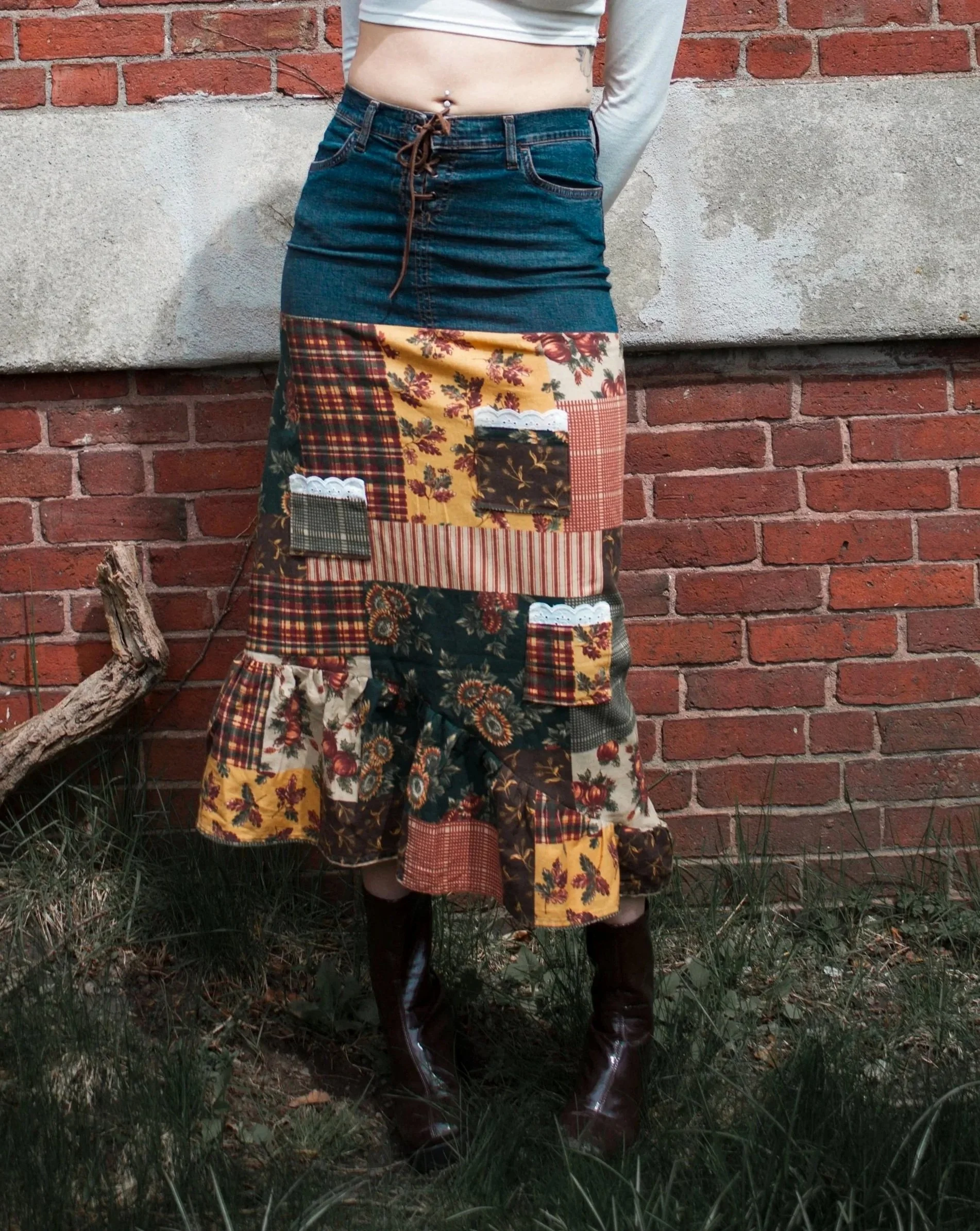 A person standing outdoors against a brick wall, wearing a long patchwork skirt with various floral and plaid patterns, a white crop top, and dark brown boots.