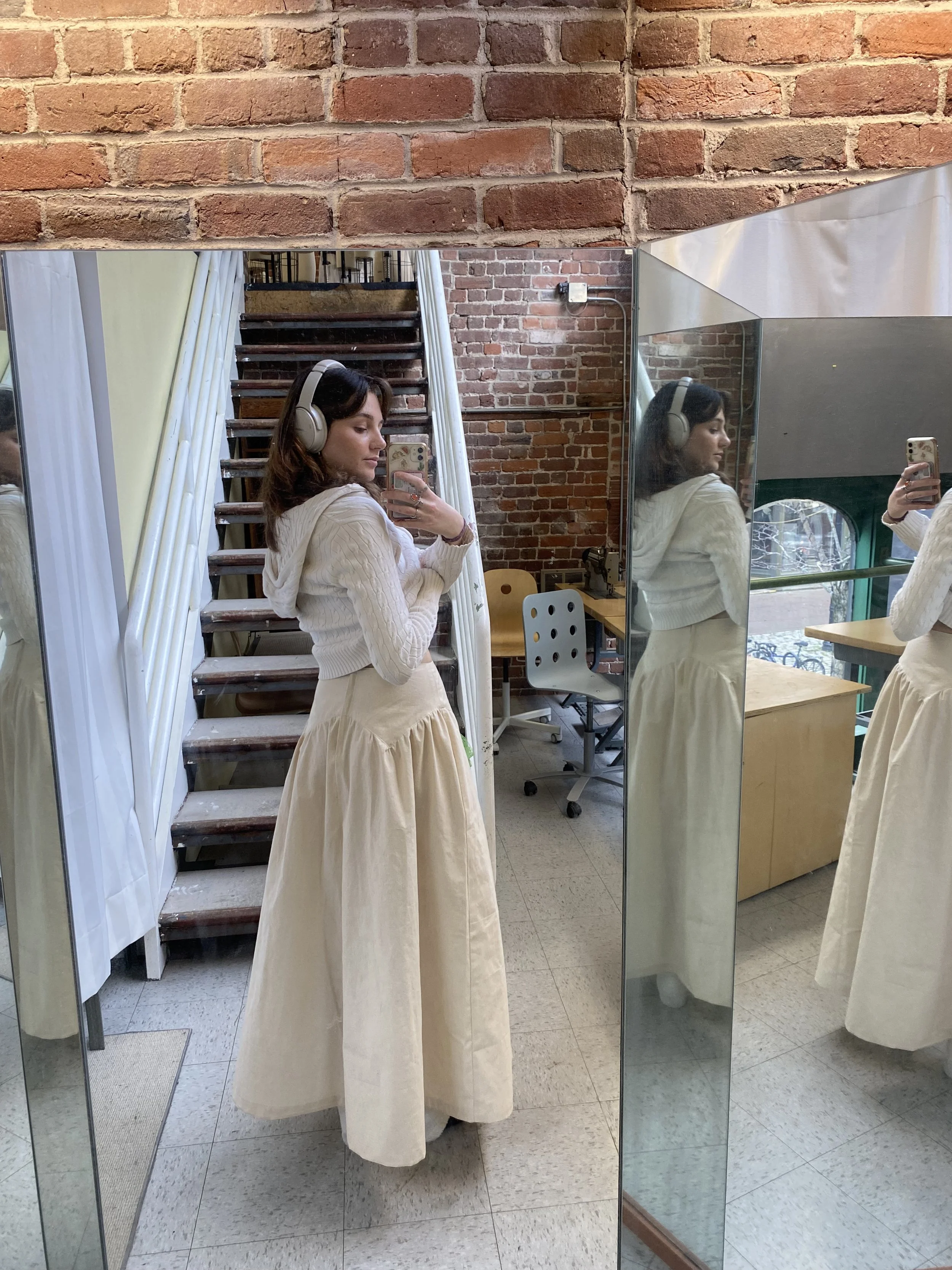 Young woman taking a mirror selfie wearing headphones, beige hoodie, and long beige skirt in an interior with brick walls and a staircase.