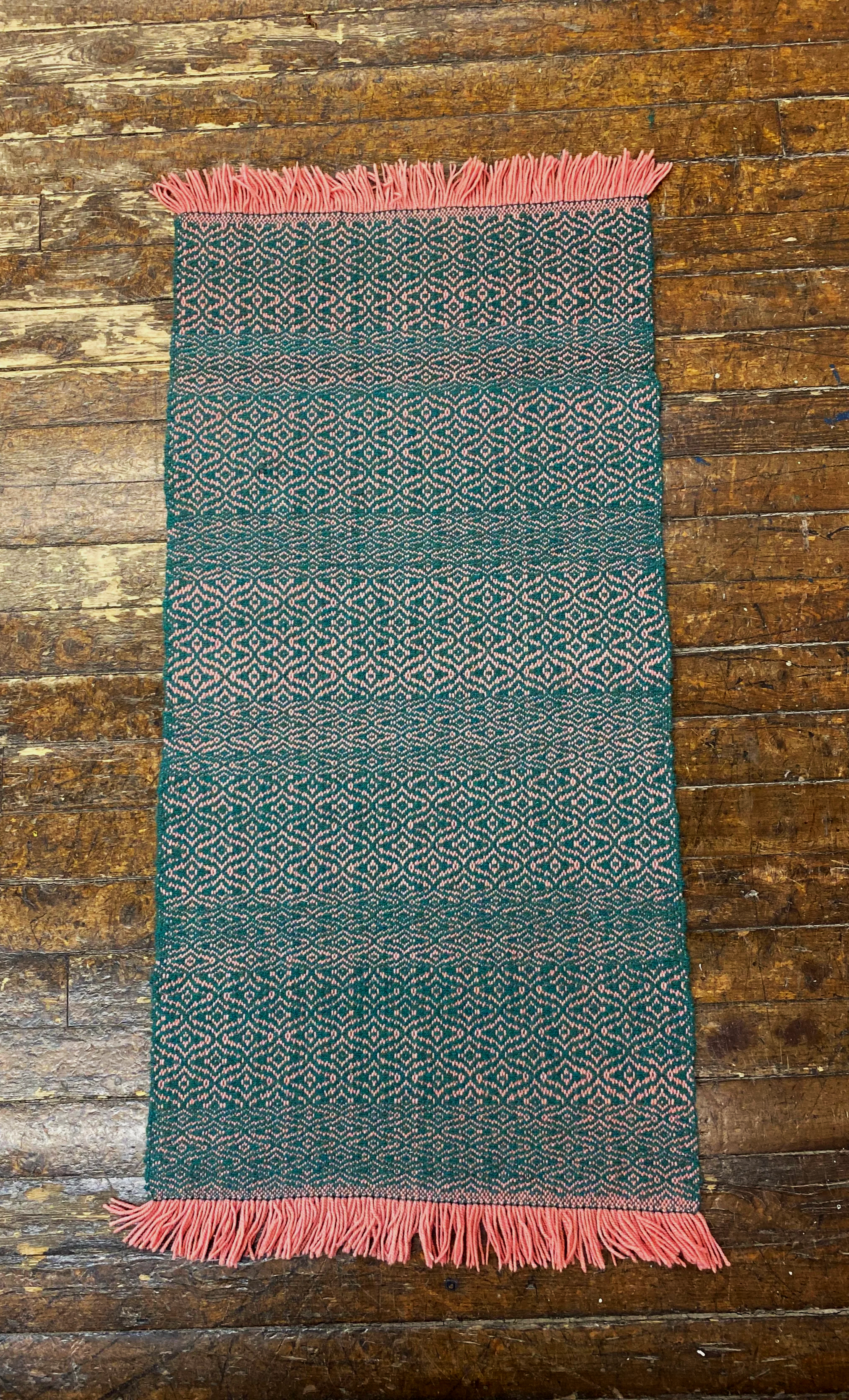 A rectangular woven rug with pink fringes on both ends, featuring a teal background with intricate pink geometric patterns, laid on a wooden floor.