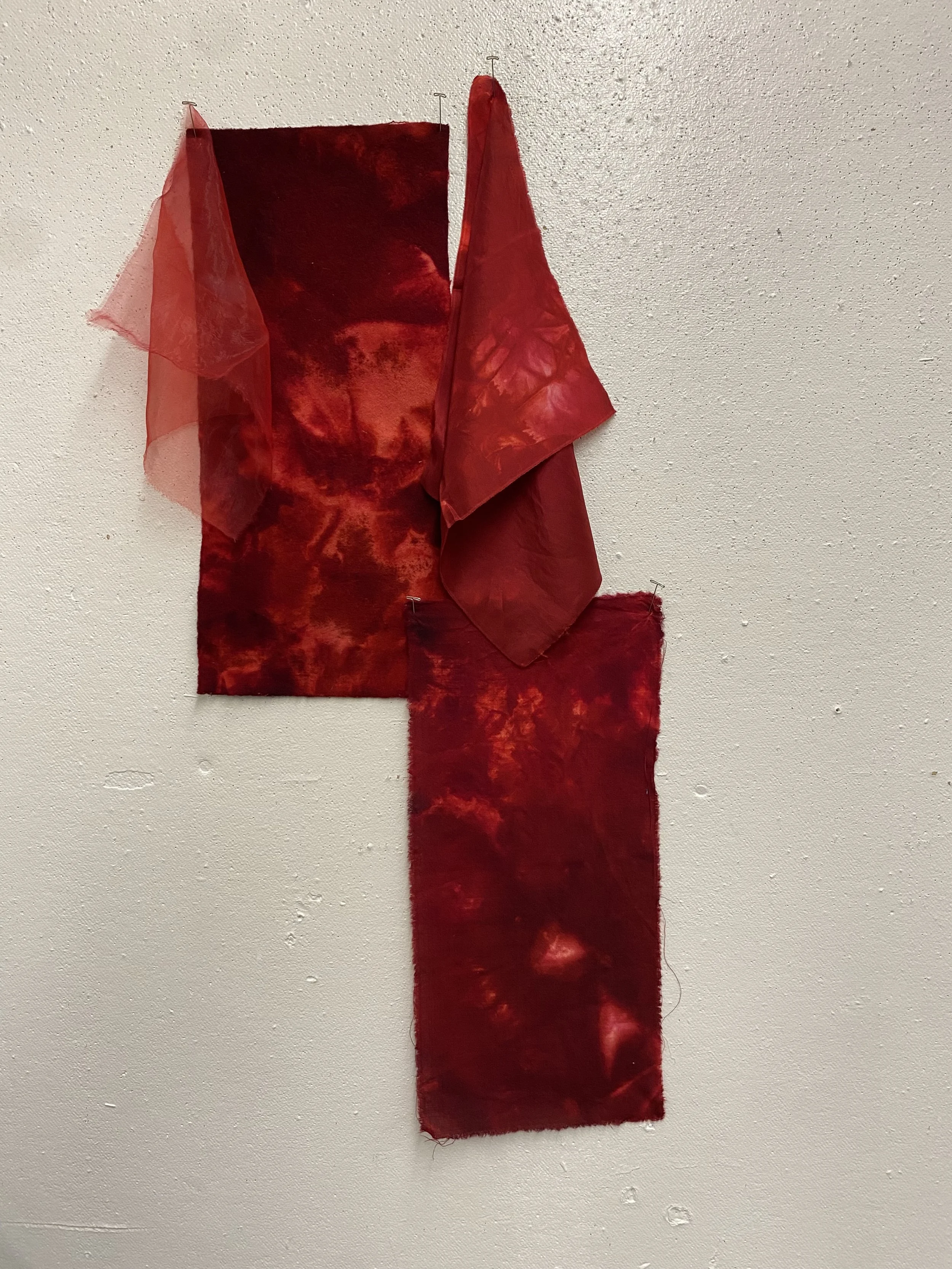 Abstract fabric art display with red and tie-dye patterns hanging on a white wall.