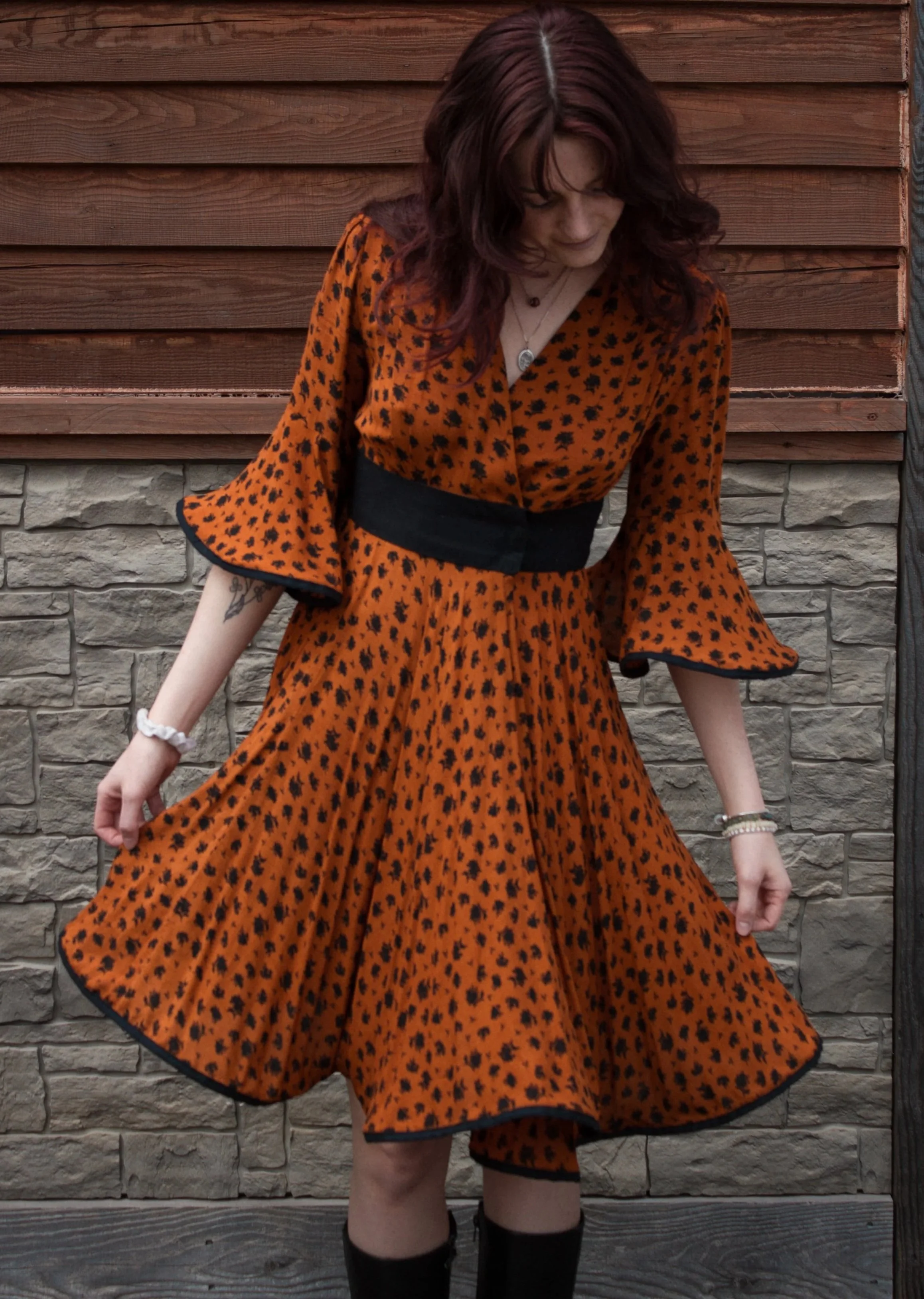 Woman in an orange and black polka dot dress standing outdoors in front of a wooden and stone wall.