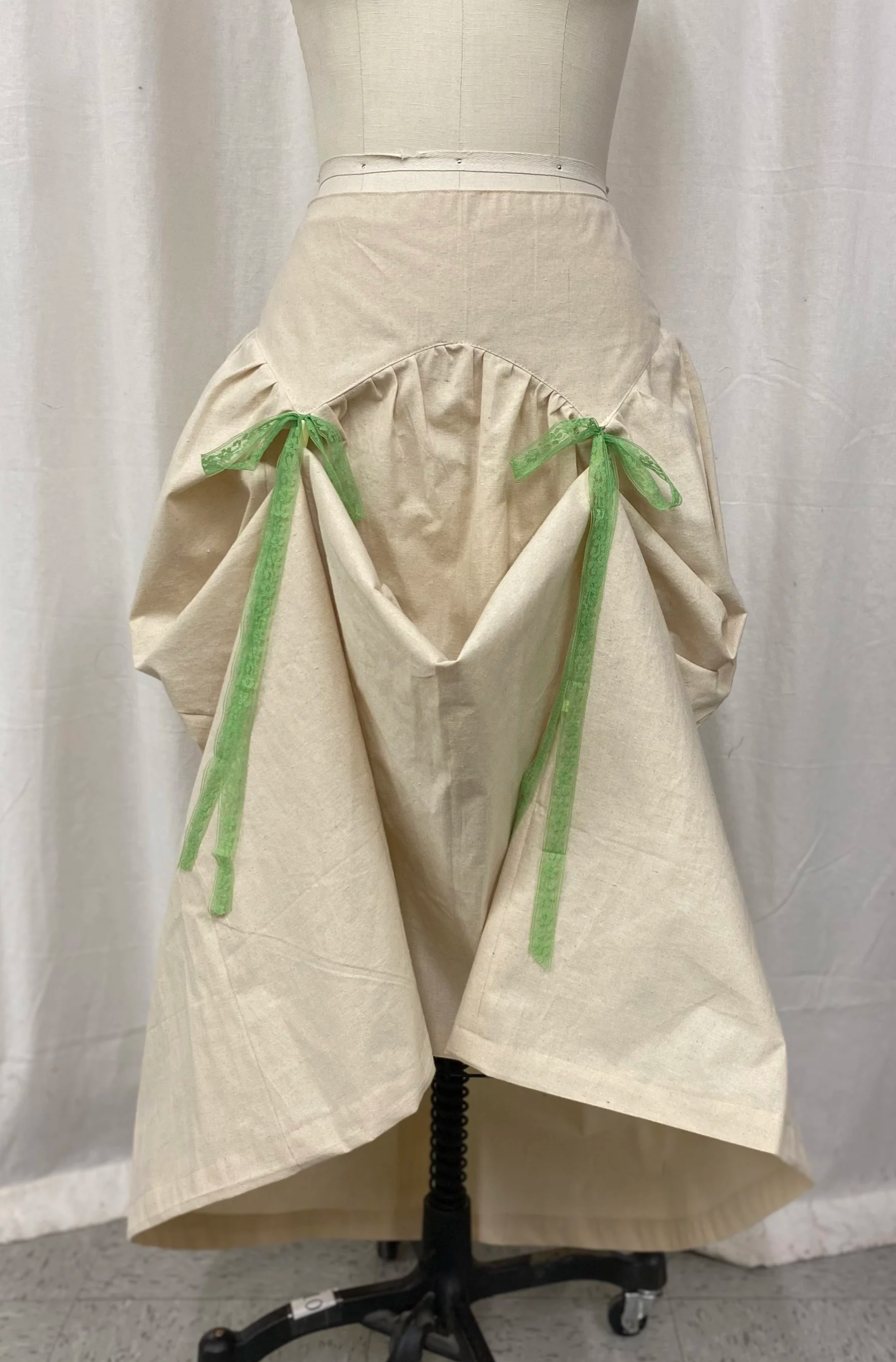 A mannequin wearing a beige skirt with green ribbon bows on the sides, against a white curtain background.
