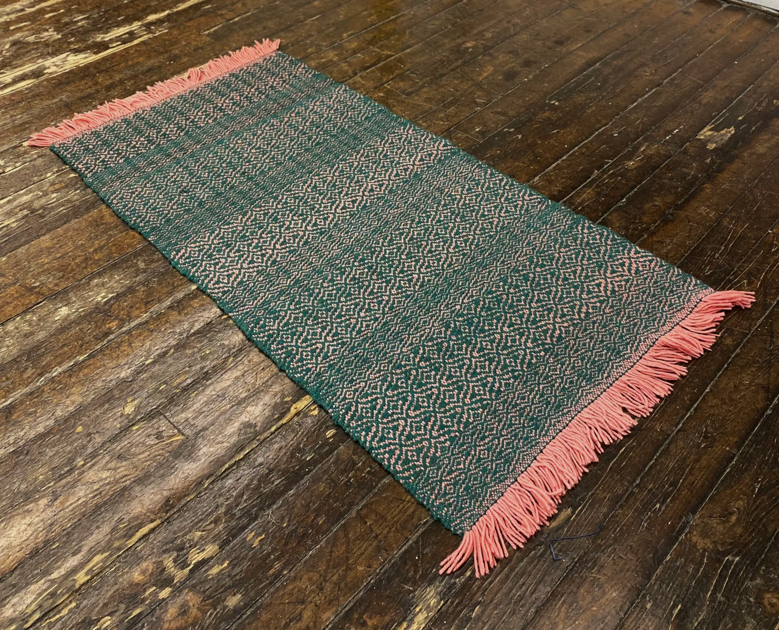 A patterned rectangular rug with pink fringes on a wooden floor.