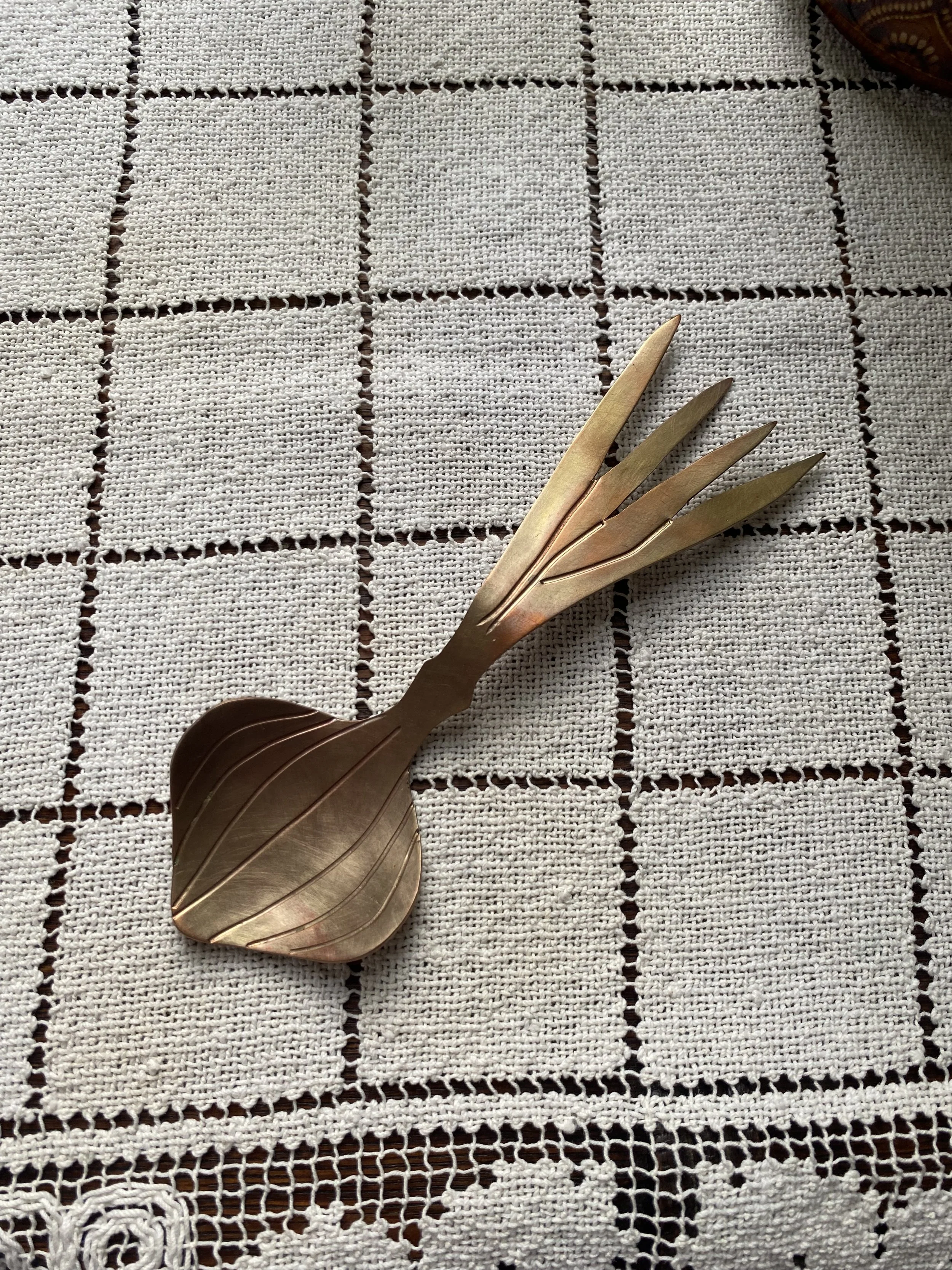 A decorative metal utensil resembling a bird with a large rounded wing and elongated beak, laid on a white crochet tablecloth with a grid pattern.