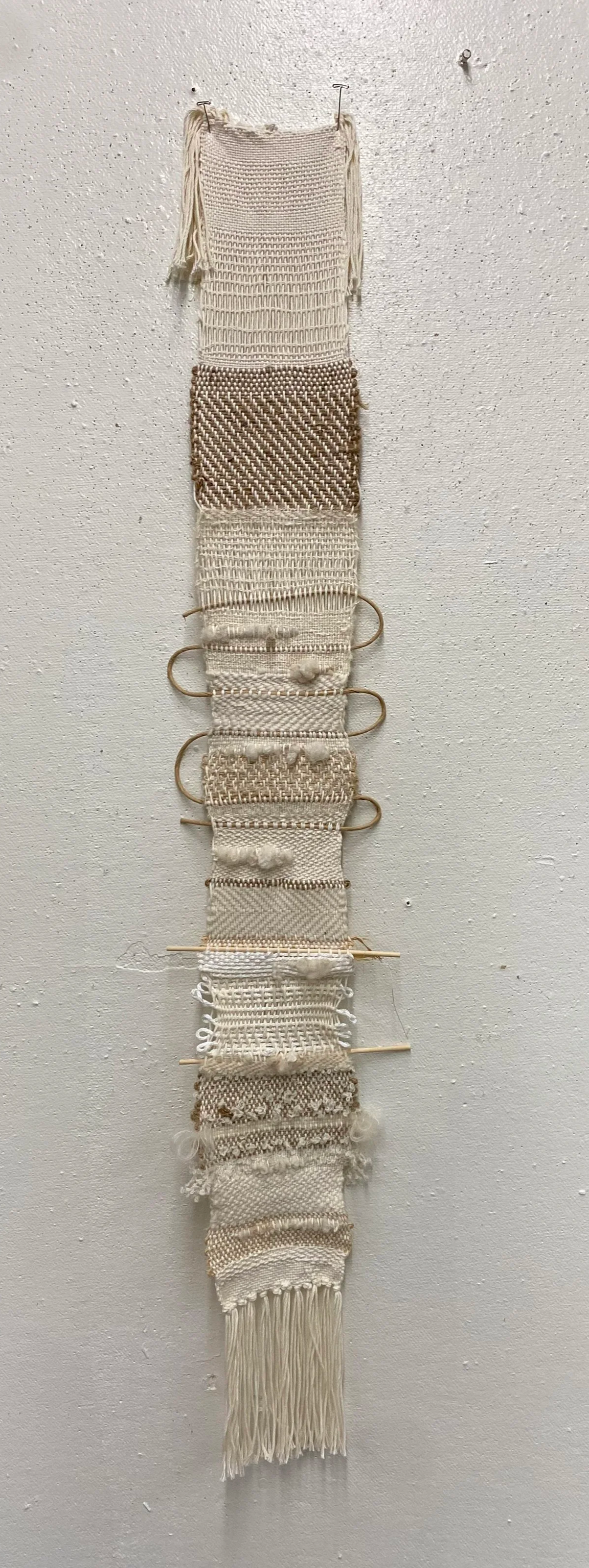 A vertical woven wall hanging made of various textured cream and beige yarns, with fringed edges at the top and bottom, hanging on a nail on a white textured wall.