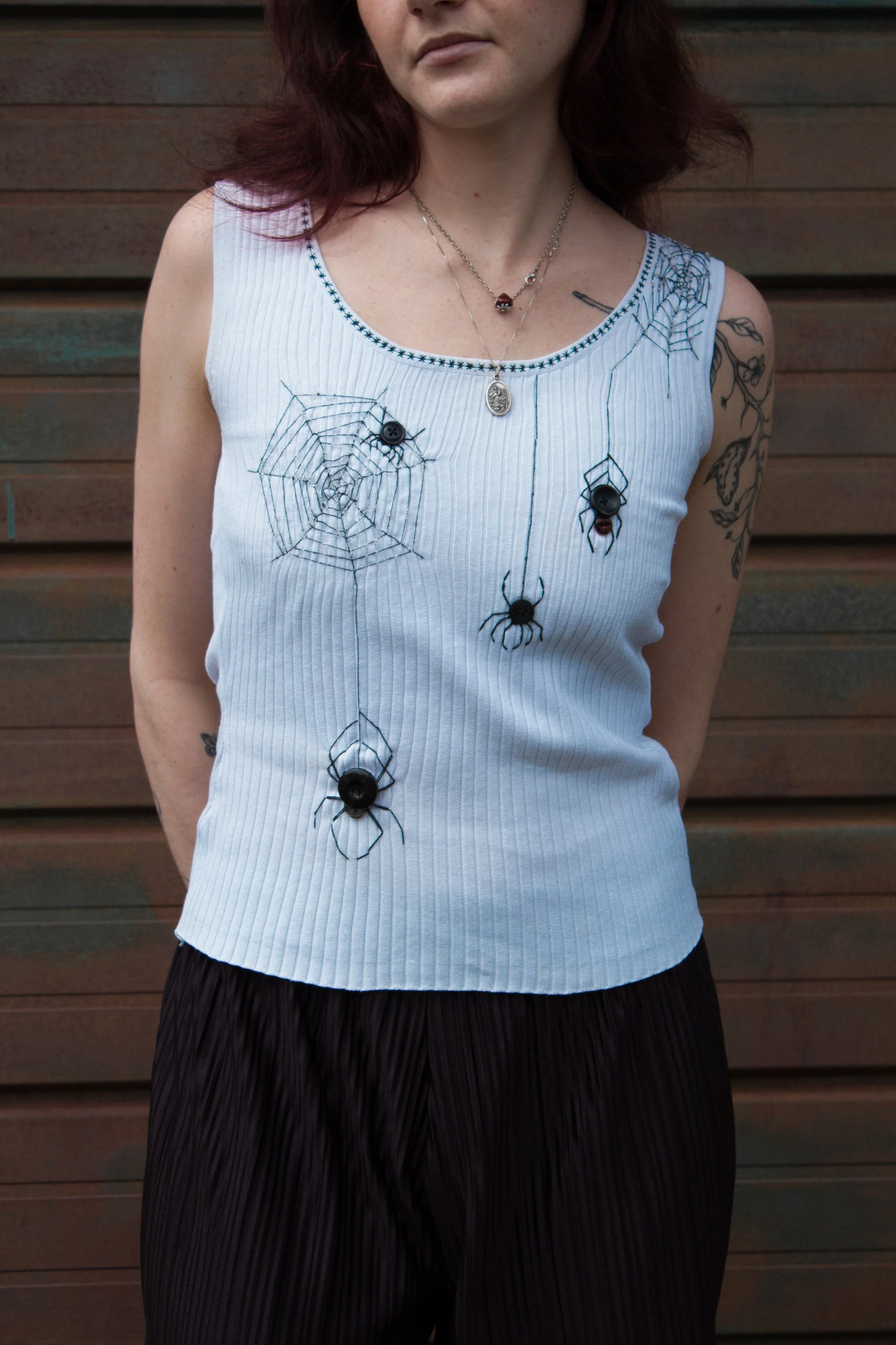 A person with red hair wearing a white sleeveless top decorated with spider, spider web, and bug appliqués, standing in front of a wooden wall.